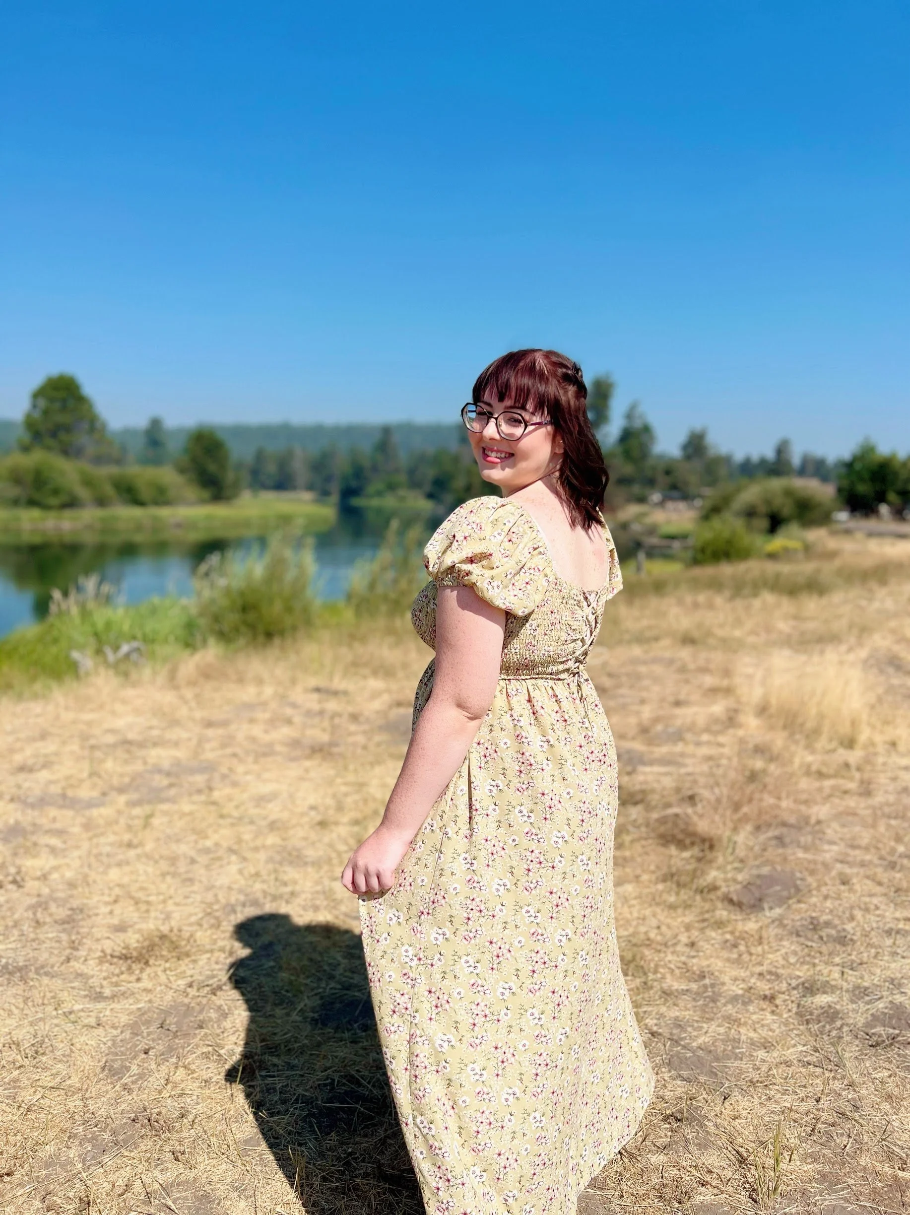 A woman with glasses and brown hair standing outdoors in a yellow floral dress, smiling, with a river and green trees in the background on a sunny day.