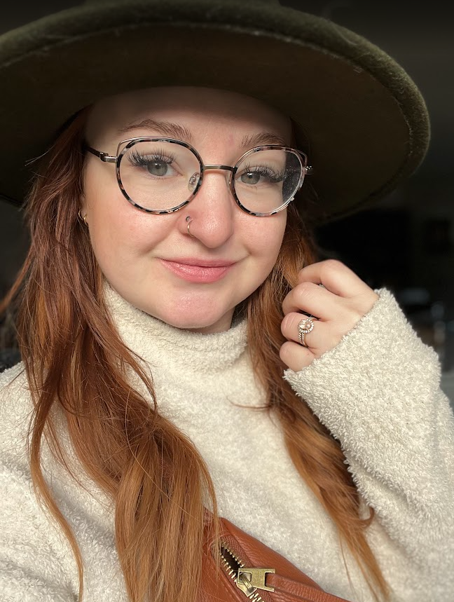 Young woman with red hair, wearing glasses, a beige turtleneck sweater, and a wide-brimmed hat, smiling and holding her hand near her face with a ring on her finger.