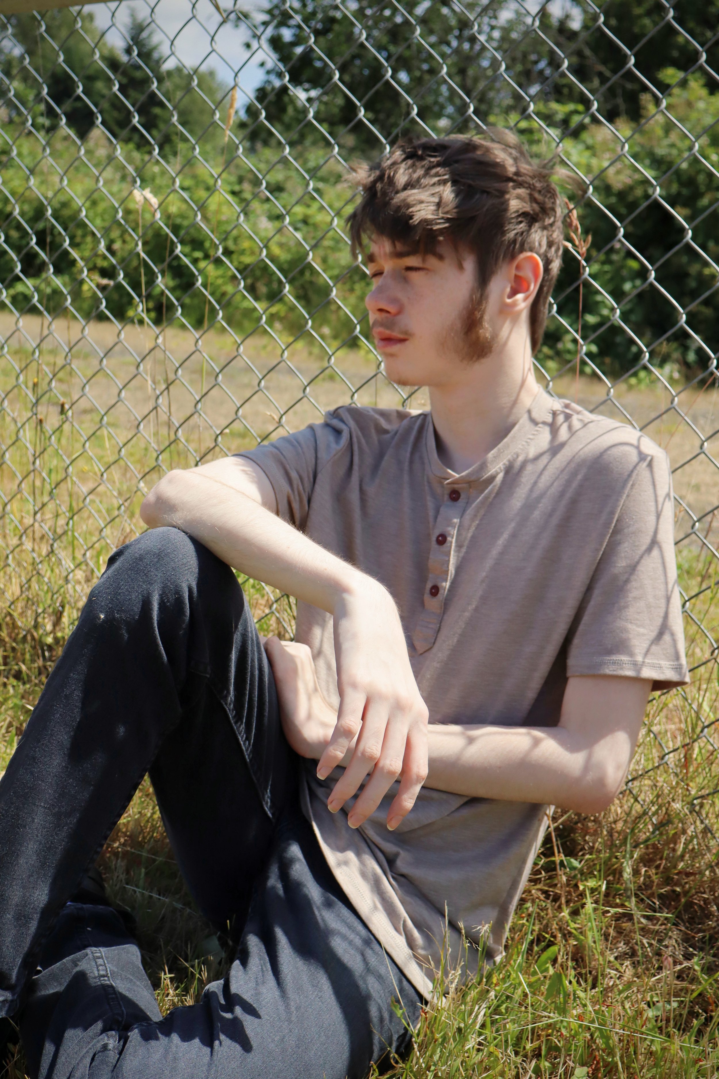 Young man with brown hair and sideburns sitting outdoors by a chain-link fence, wearing a beige shirt and black pants, with trees and grass in the background.