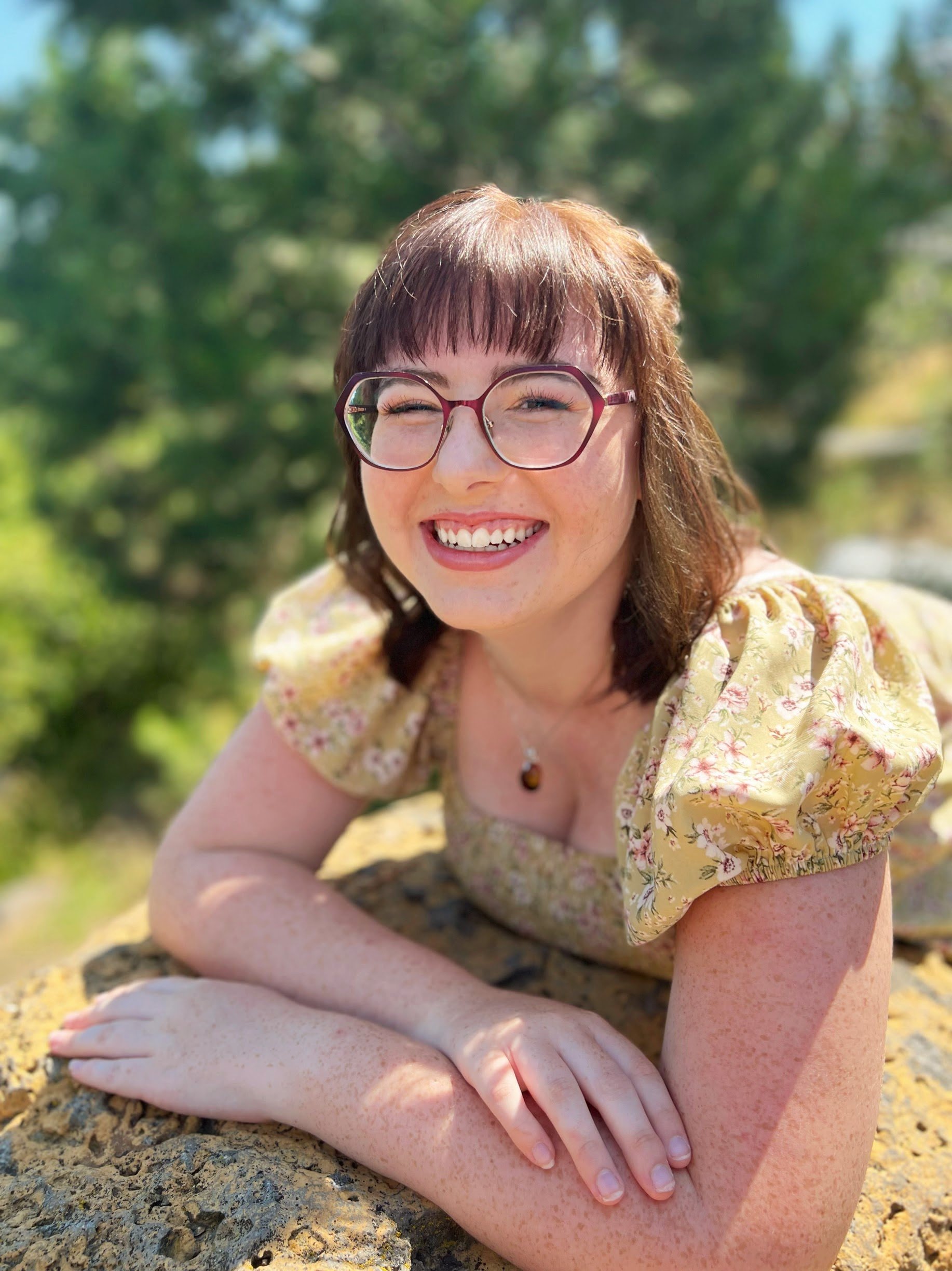 A woman with glasses and a yellow floral dress crouching outdoors in sunlight, smiling with closed eyes, on a rocky surface with green trees in the background.