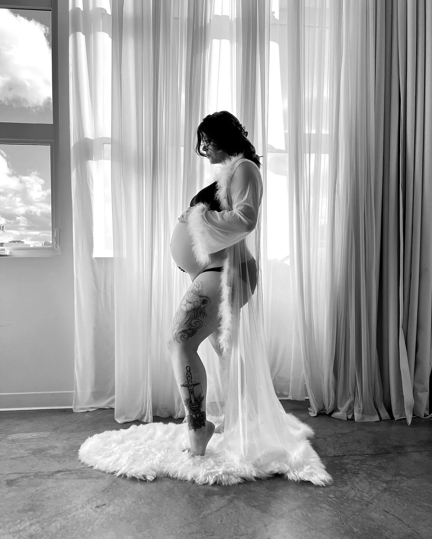 A pregnant woman with tattoos on her legs standing on a fluffy rug in front of sheer curtains, with a window showing a cloudy sky in black and white.