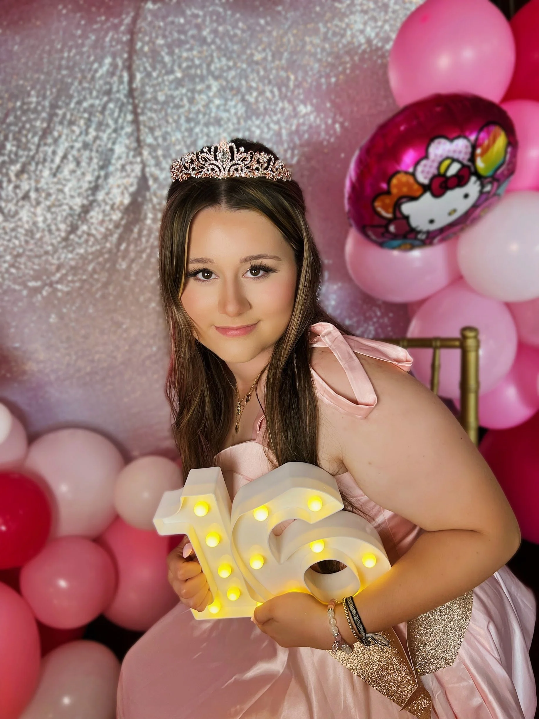 A young woman at a birthday party, wearing a tiara and holding a lighted decorative number '16' sign, surrounded by pink and white balloons, with a pink glittery backdrop.