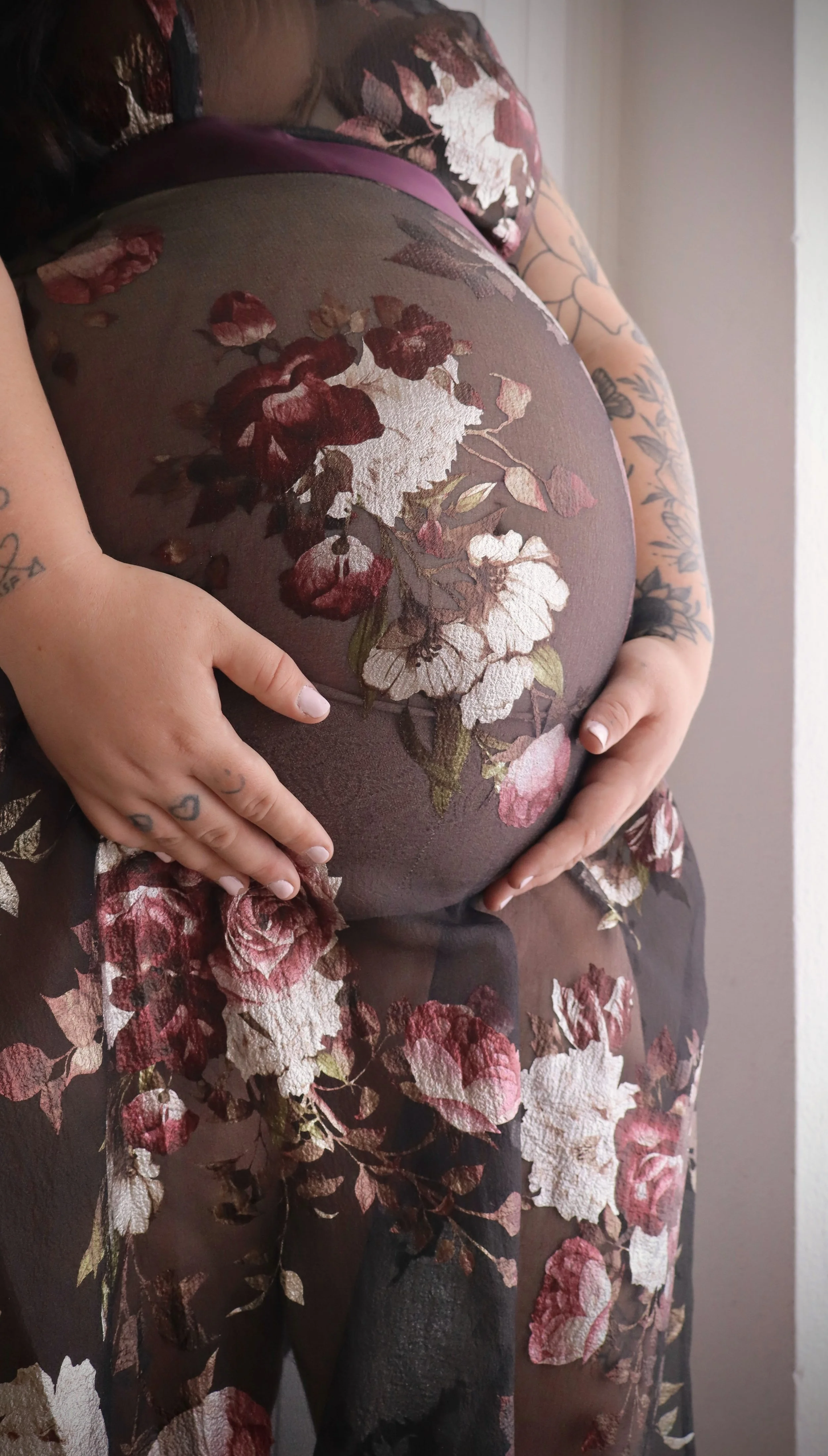 A pregnant woman wearing sheer floral black fabric with pink and cream flowers, gently holding her belly with both hands.