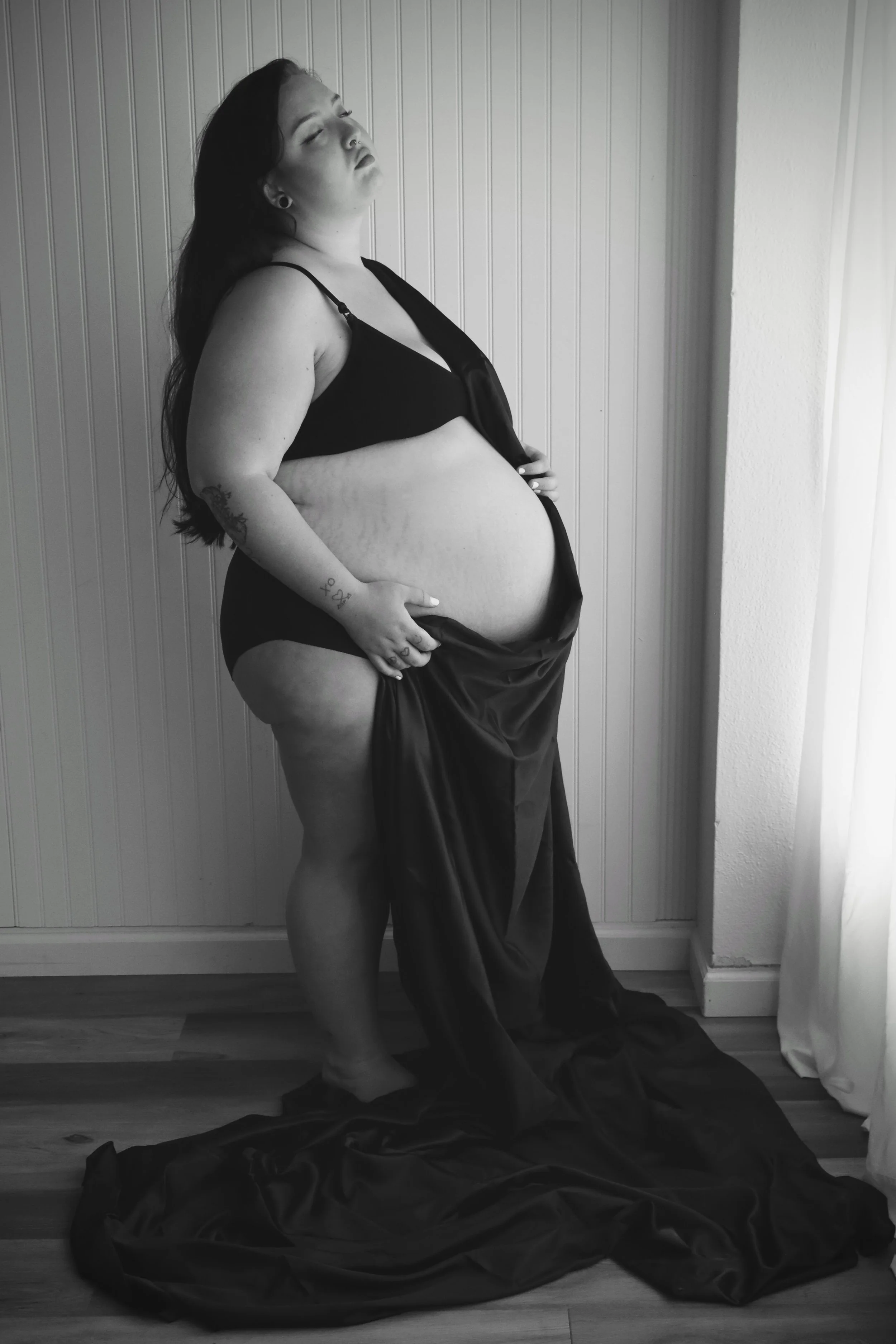 A woman with long dark hair, a nose piercing, and tattoos on her arm, stands against a wall with vertical wooden paneling. She wears black lingerie and a satin robe, with her eyes closed and her head tilted back slightly. She holds her abdomen with one hand, and a dark fabric drapes down to the floor, pooling around her feet. The photo is in black and white.