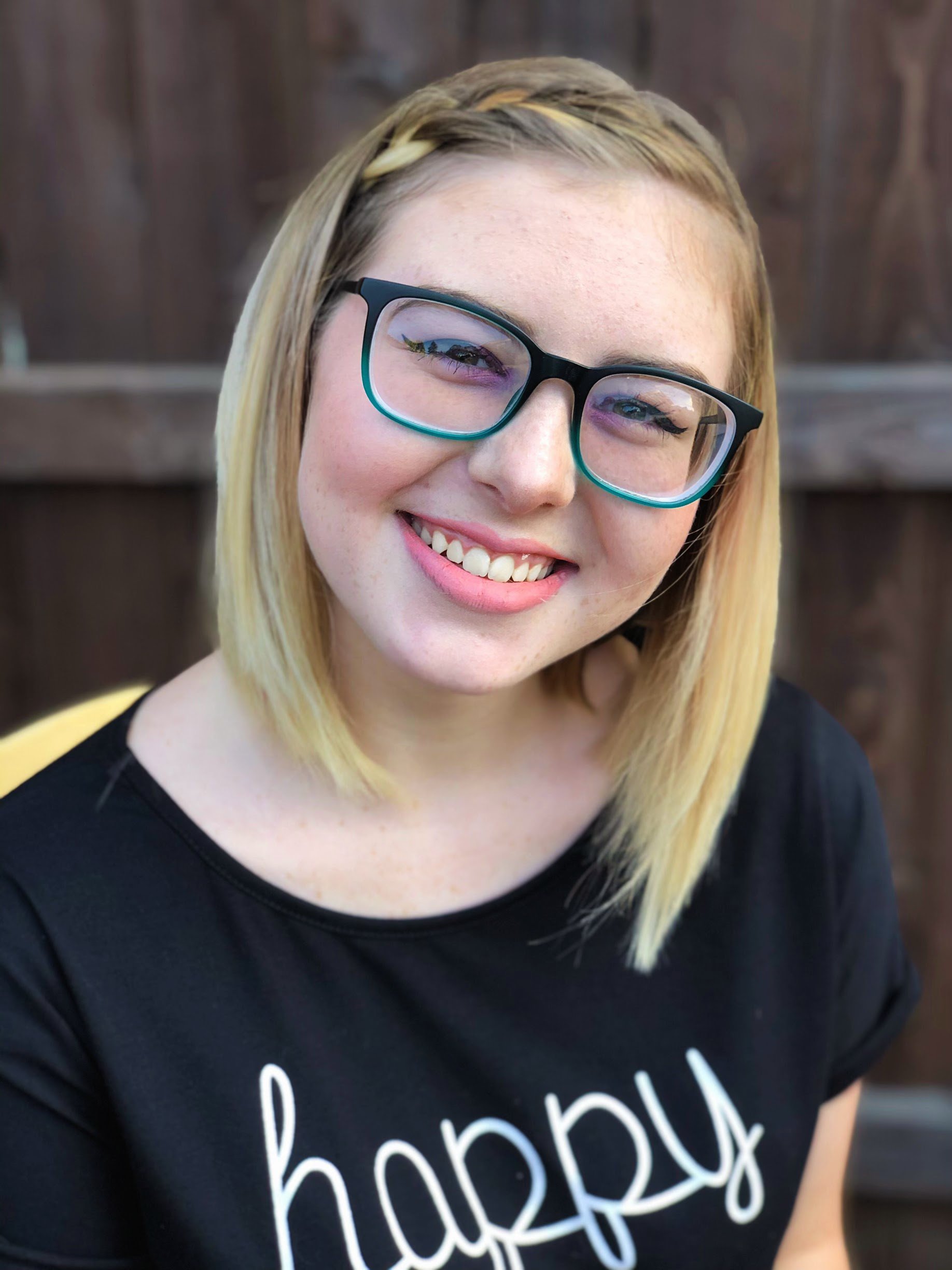 Close-up of a smiling woman with blonde hair and black-rimmed glasses, wearing a black shirt with white letters, standing outdoors with a wooden fence background.