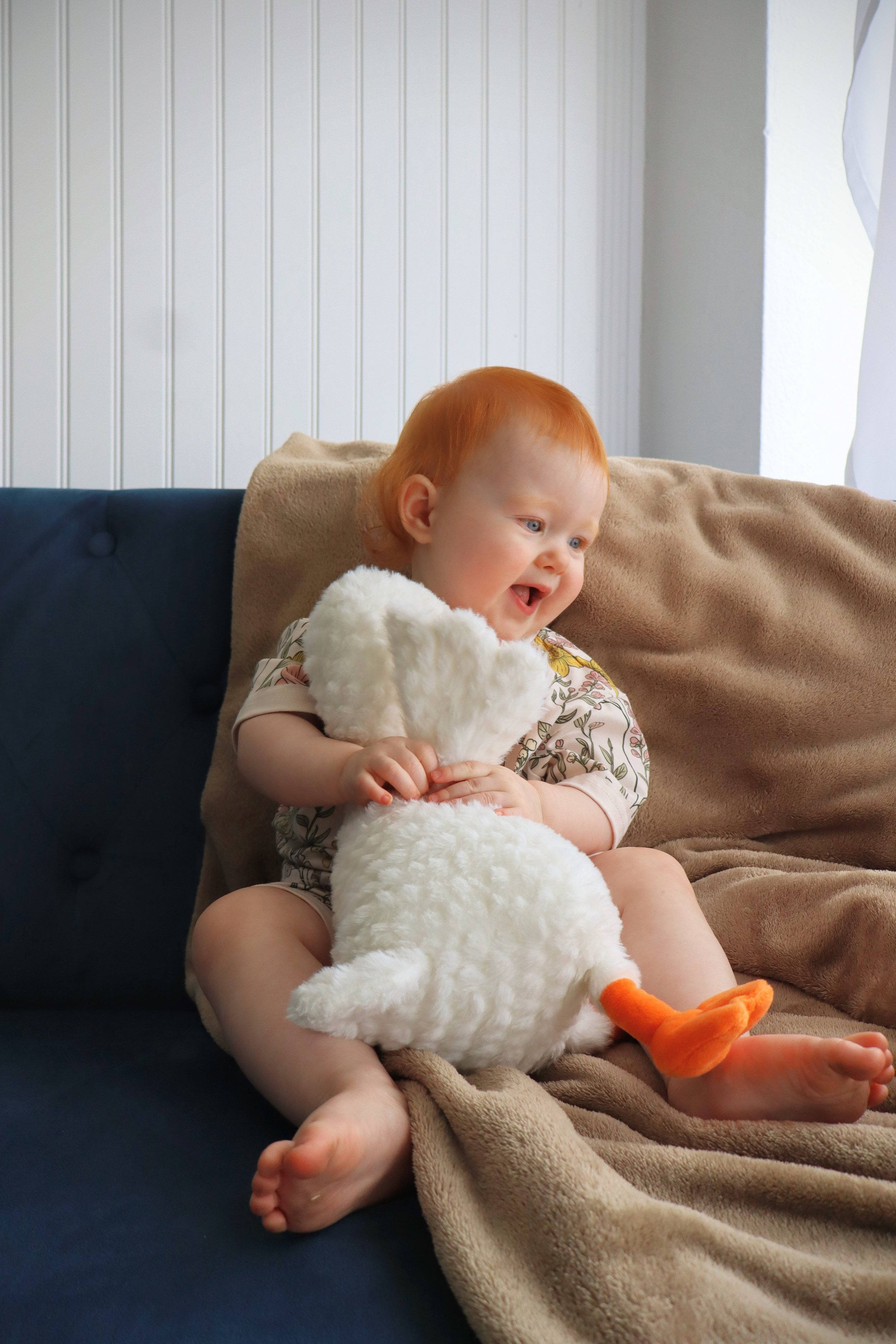 A young child with red hair sitting on a couch, holding a plush duck stuffed animal, and wearing a floral shirt and shorts. The child is smiling and looking to the side. The background features a white paneled wall and a beige blanket draped over the couch.