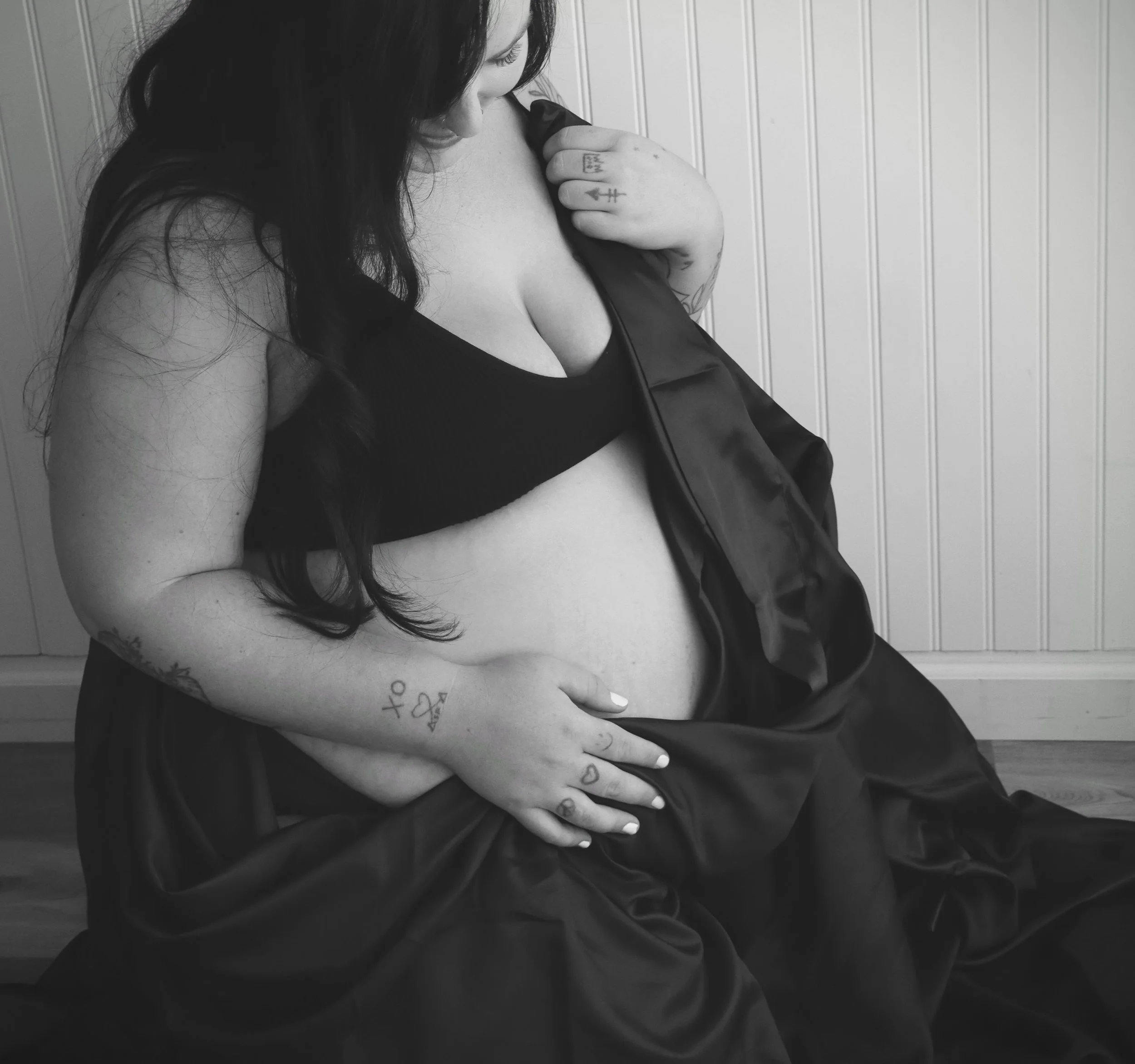 Black and white photo of a woman with long dark hair wearing a black bra, holding her shoulder with her right hand that has tattoos. She is looking downward, with a white paneled wall in the background.