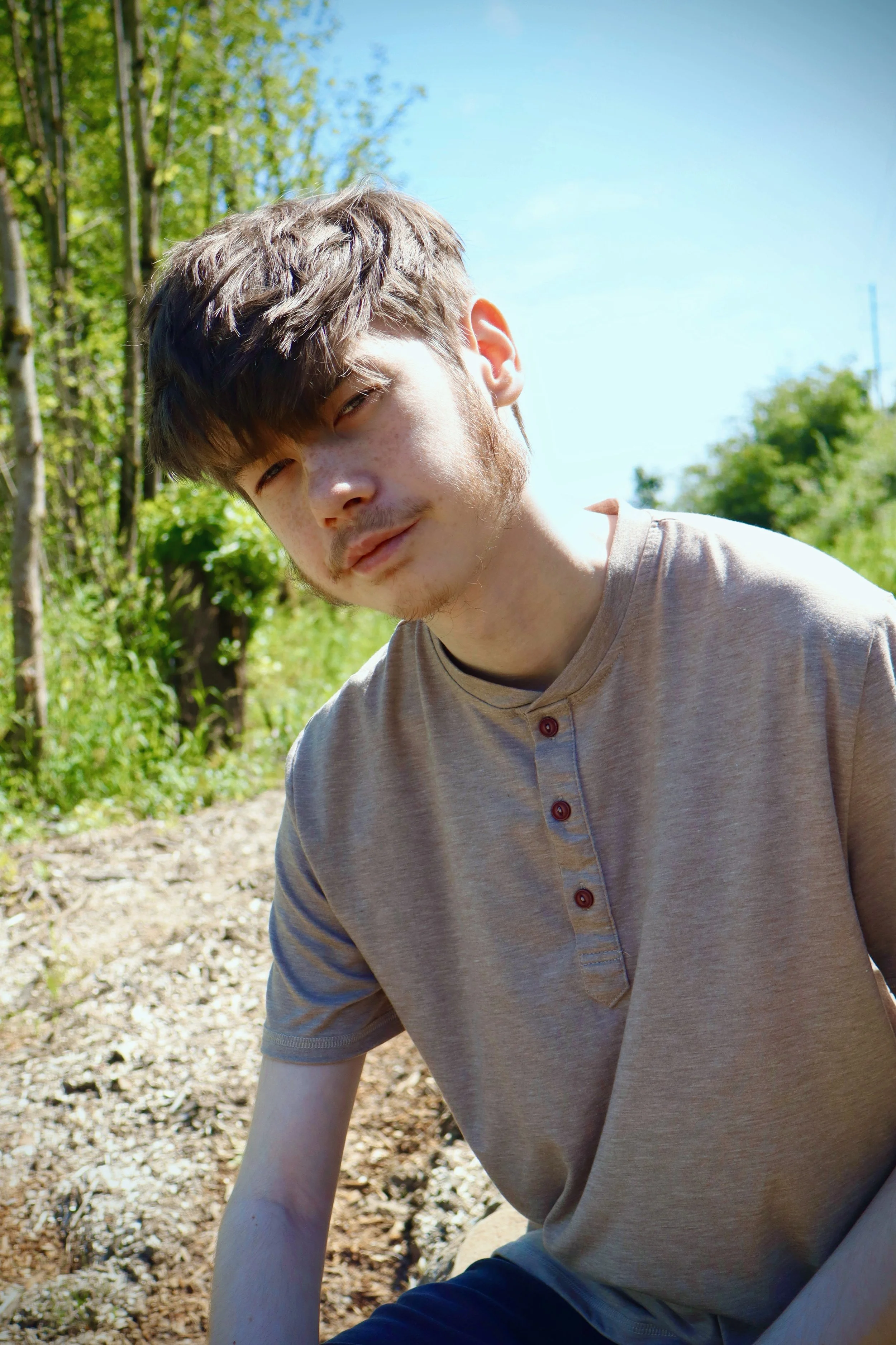 Close-up of a young man with light brown hair and freckles, sitting outdoors on a sunny day, with green trees and a blue sky in the background.