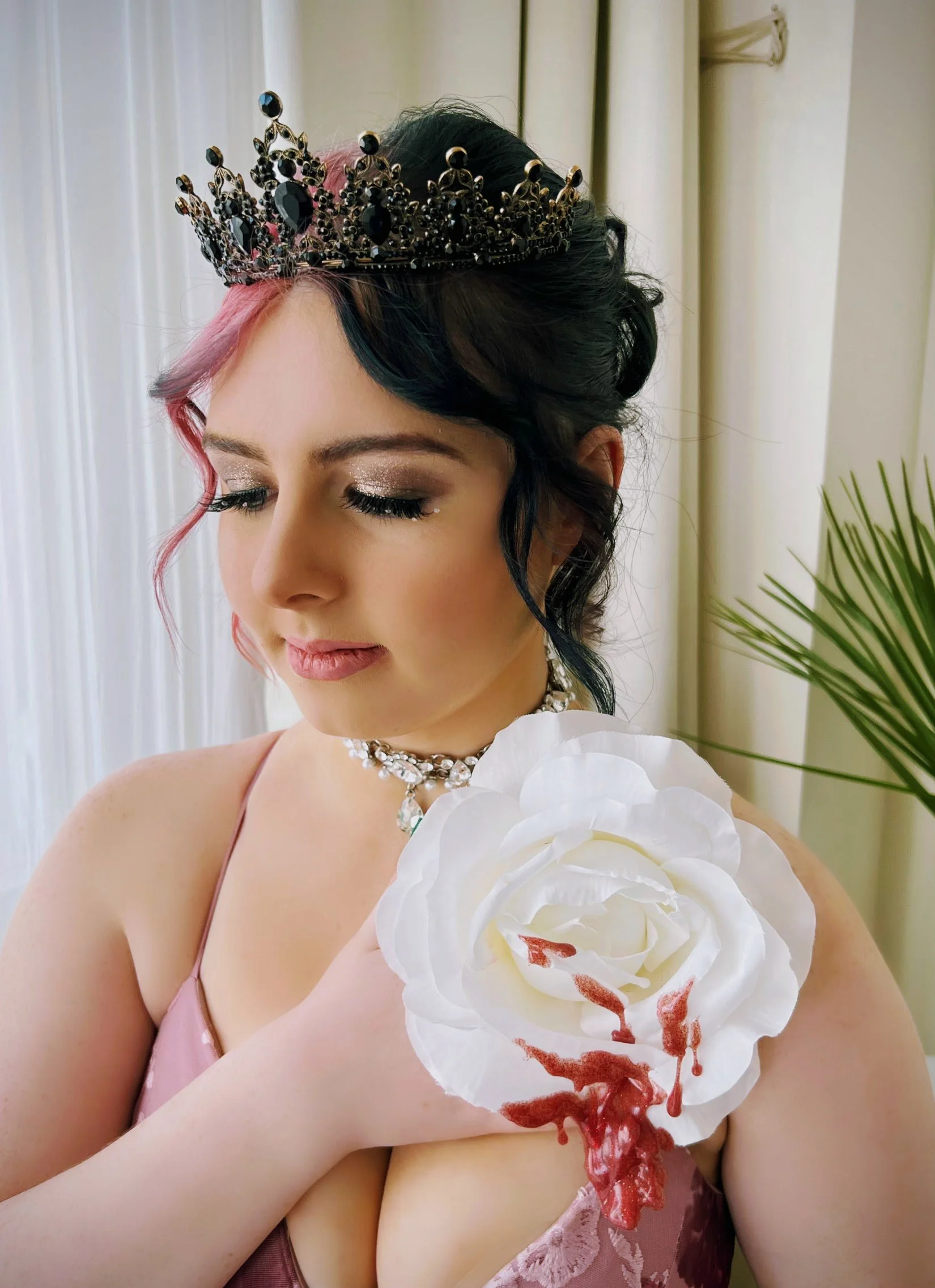 Woman wearing a crown and jewelry, holding a white flower with red makeup or paint on it, in front of a curtain and a green plant.