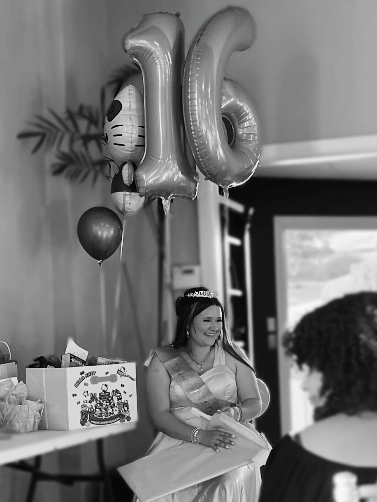 A celebration scene with a woman wearing a crown and sash, sitting with a gift bag and surrounded by balloons, indicating a birthday or special occasion.