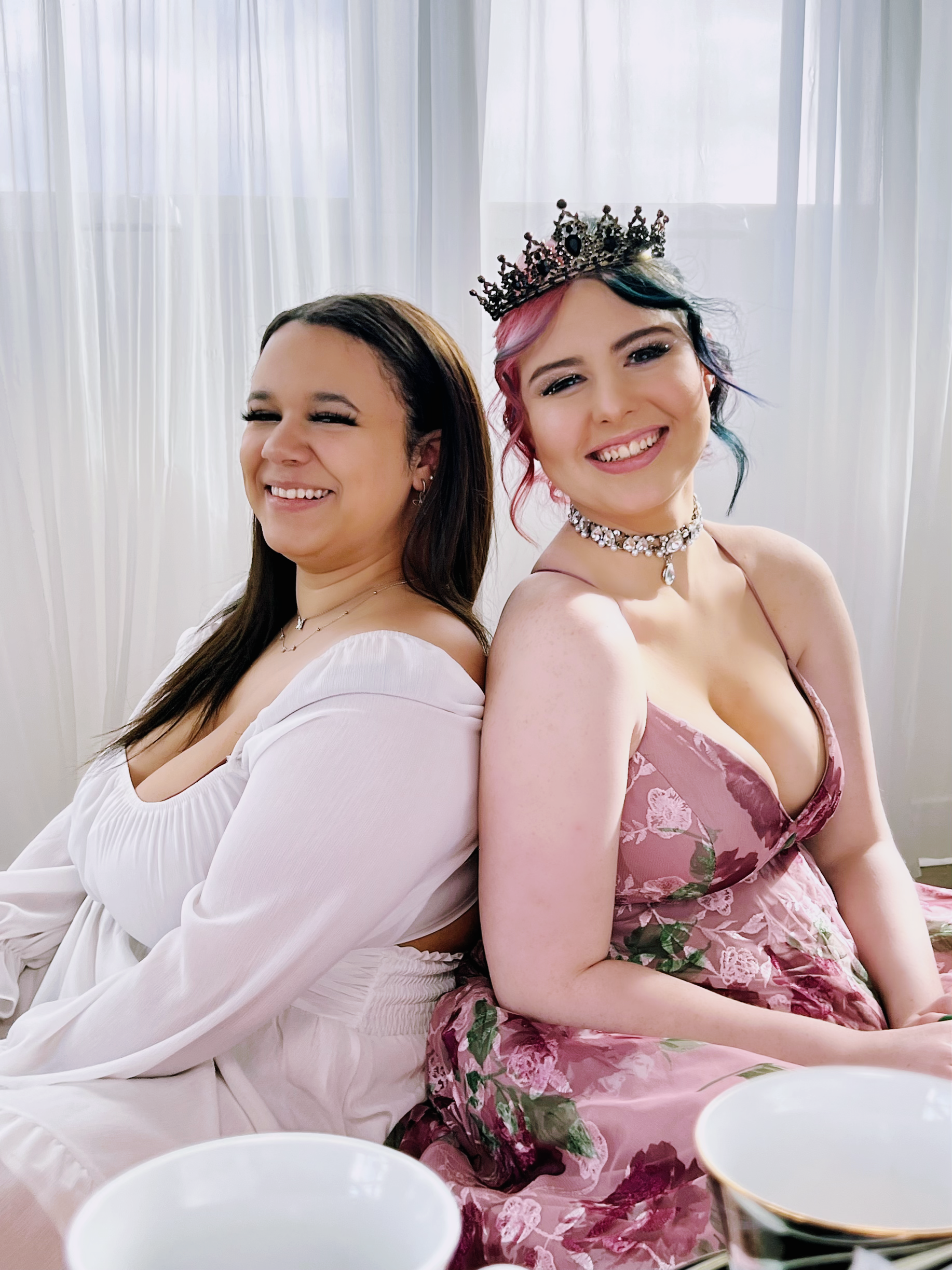 Two women sitting back-to-back smiling; one wearing a crown, jewelry, and a pink floral dress, and the other in a white dress, in front of sheer white curtains, with cups in the foreground.