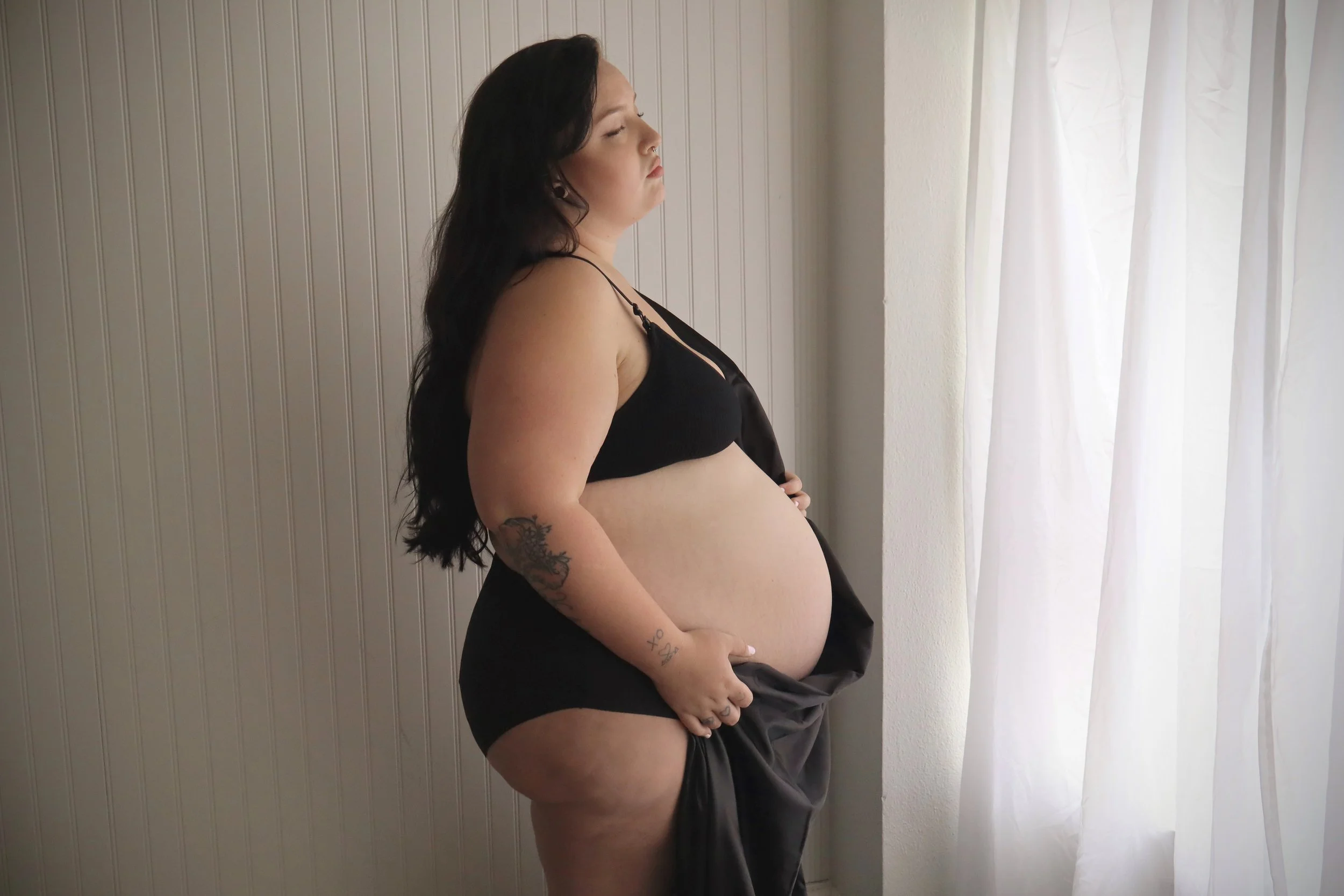 A pregnant woman with dark hair, tattoos on her arm, wearing a black bra and black shorts, stands sideways near a window with white curtains and wood-paneled wall, with her eyes closed and hand on her belly.