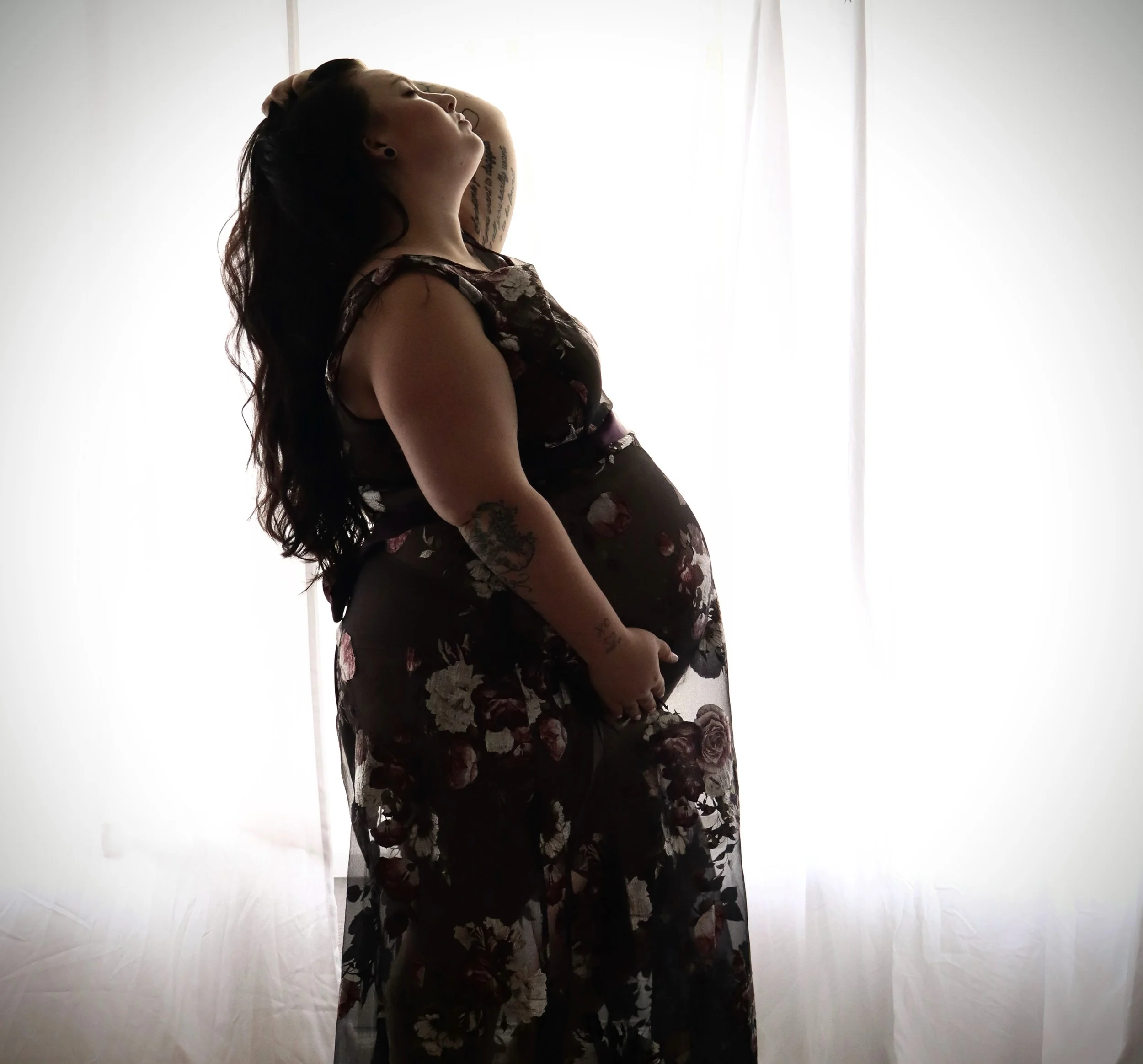 Side view of a woman with long dark hair, tattooed arms, wearing a floral dress, standing in front of a backlit white curtain with daylight coming through.