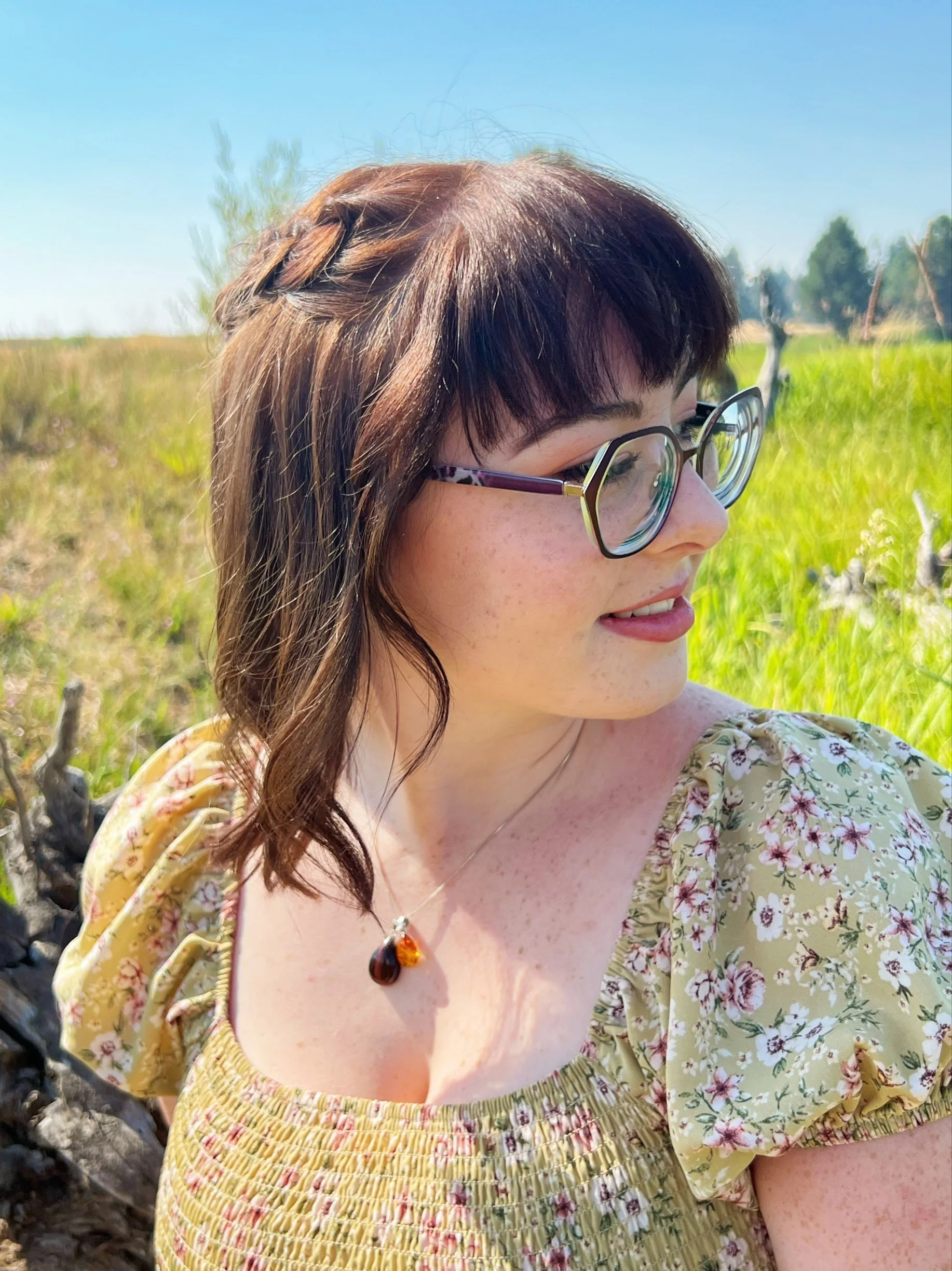 A woman with brown hair and glasses wearing a yellow floral dress and a amber jewelry pendant, outdoors in a grassy area under a bright blue sky.