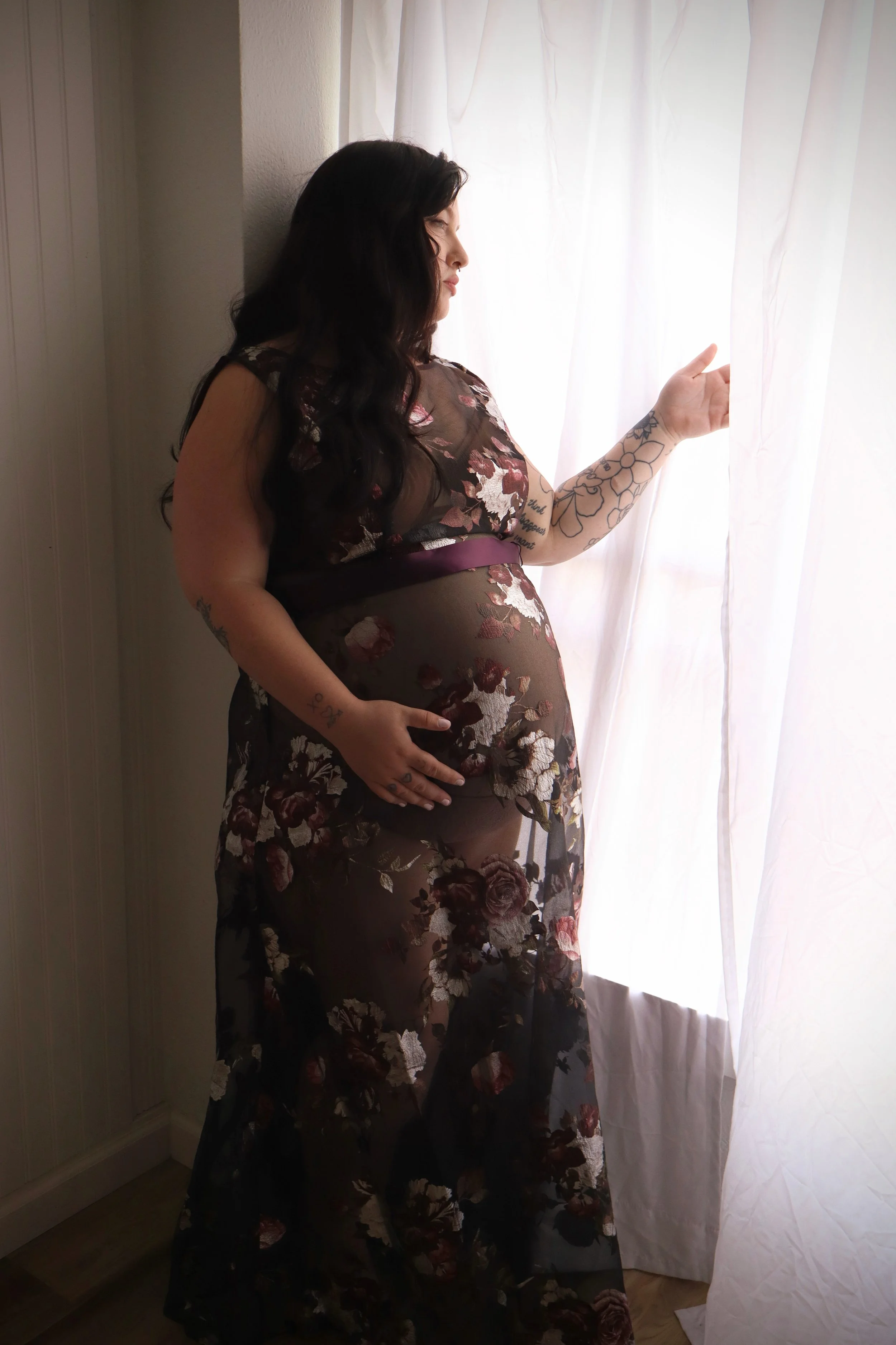 A pregnant woman with long dark hair and floral tattoos on her arms, wearing a sheer floral dress, standing by a window with white curtains, gently holding the curtain and looking out.
