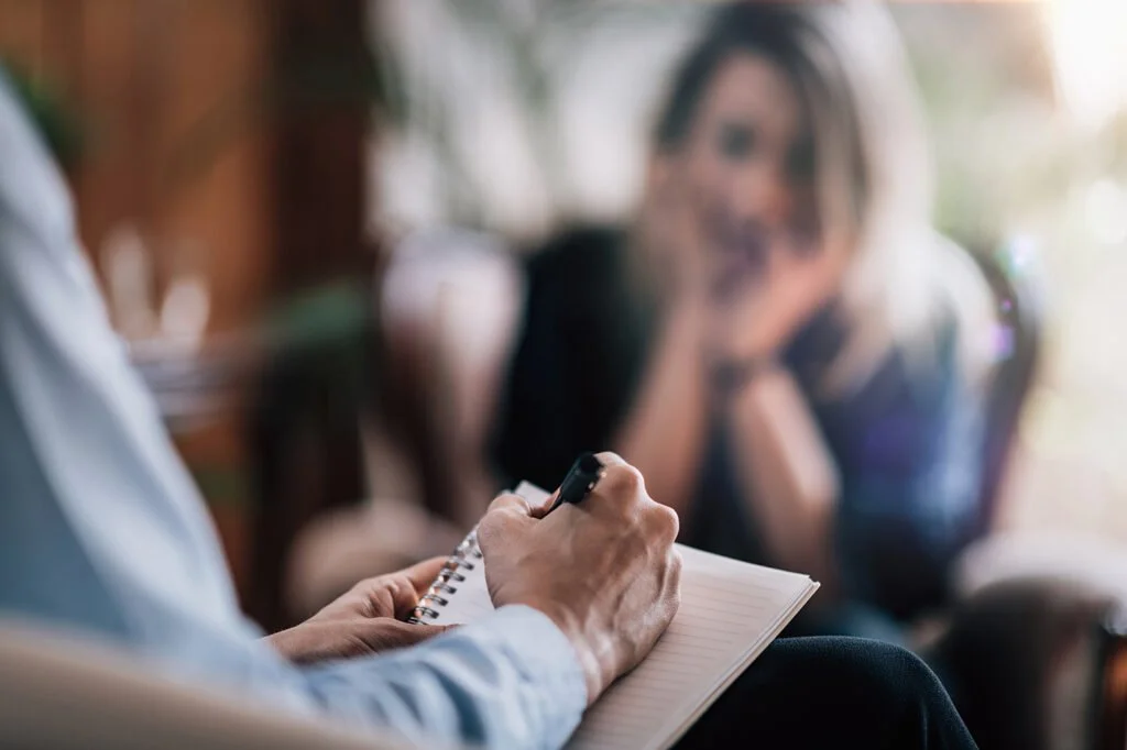 A woman sitting on a chair with her hands on her face during a therapy session or counseling, while a therapist writes notes on a notepad.