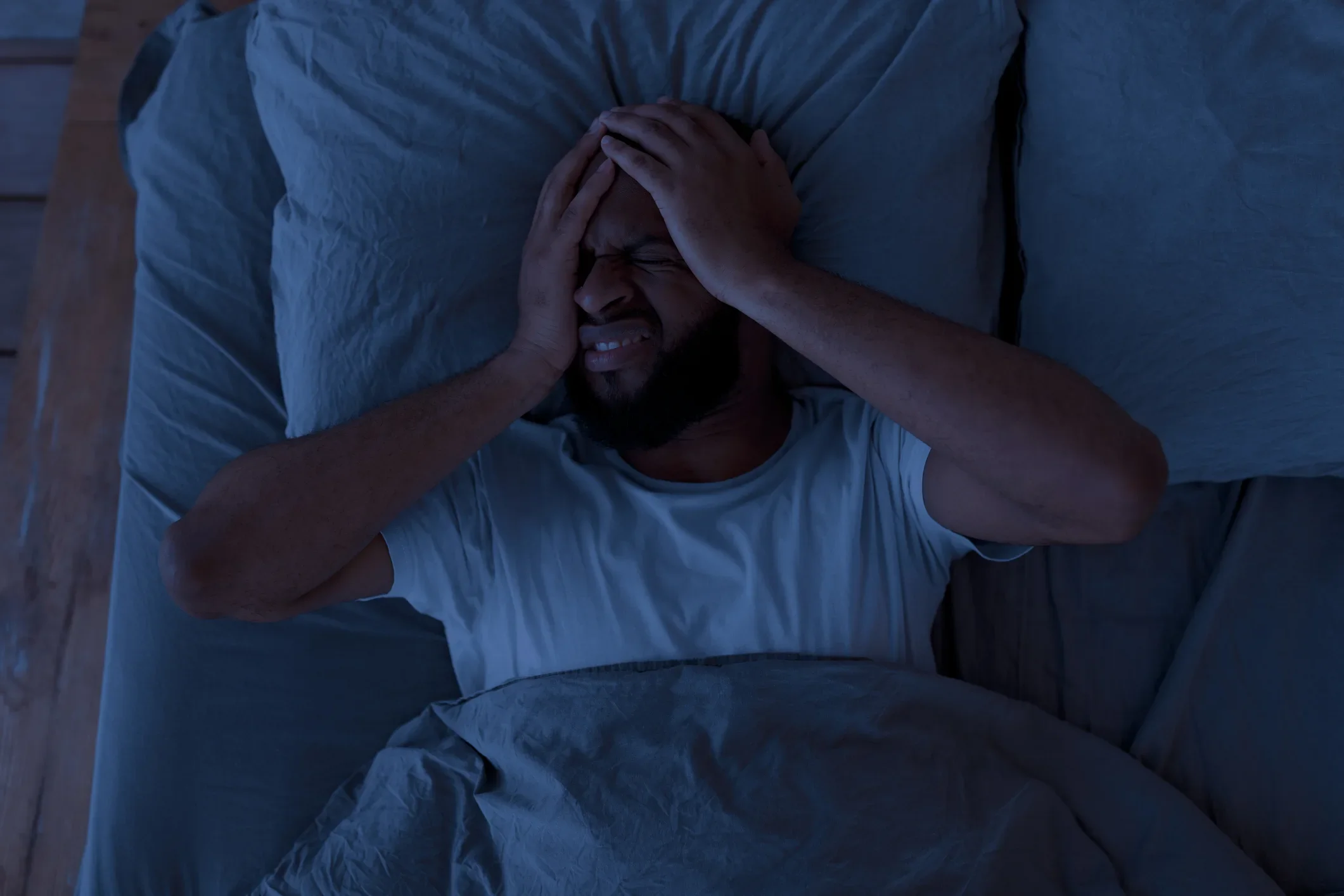A man lying in bed with eyes closed, holding his head with his hands, appearing distressed or in pain.