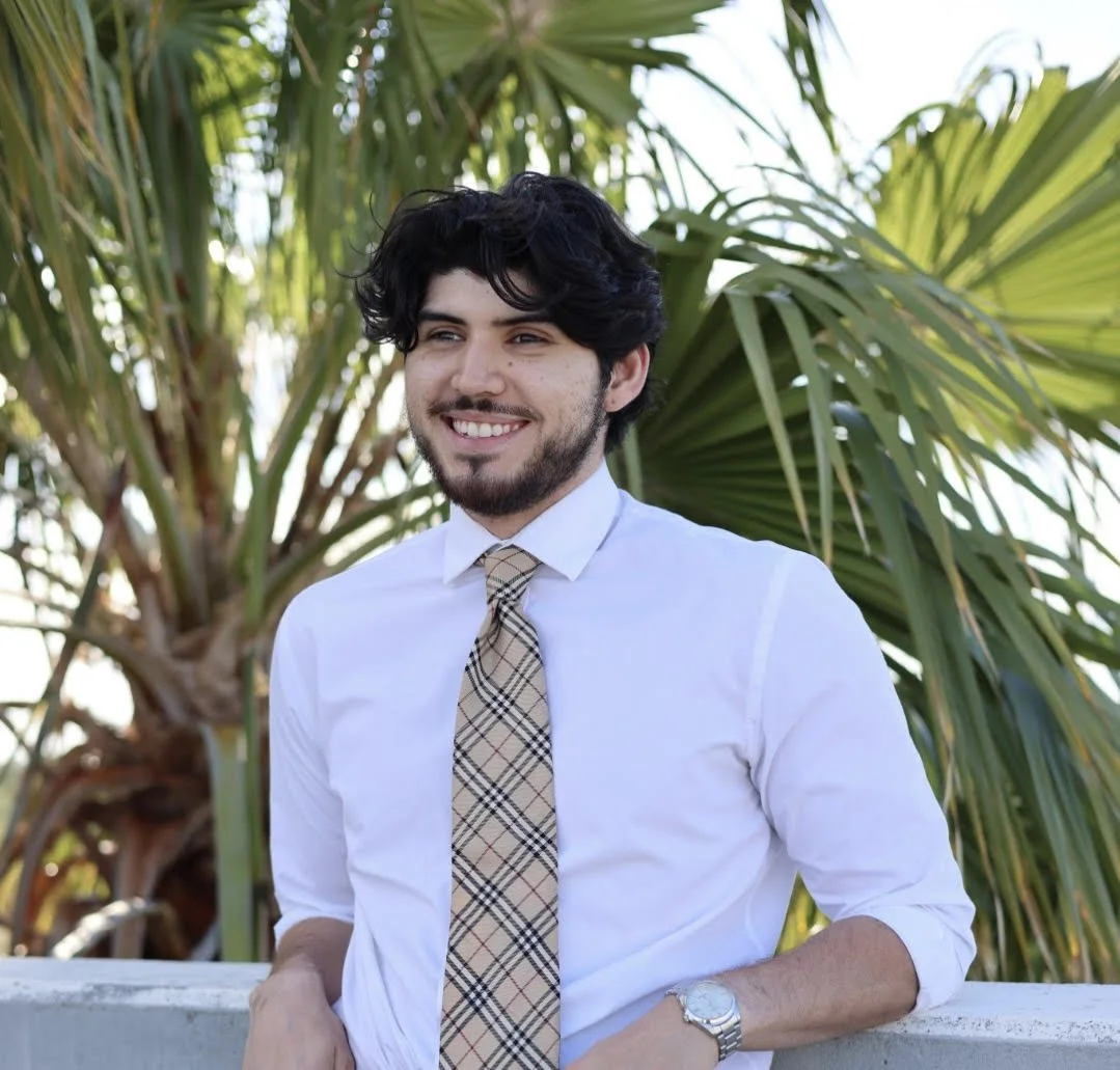 A man with dark wavy hair and a beard wearing a white shirt and patterned tie, smiling outdoors in front of green foliage.