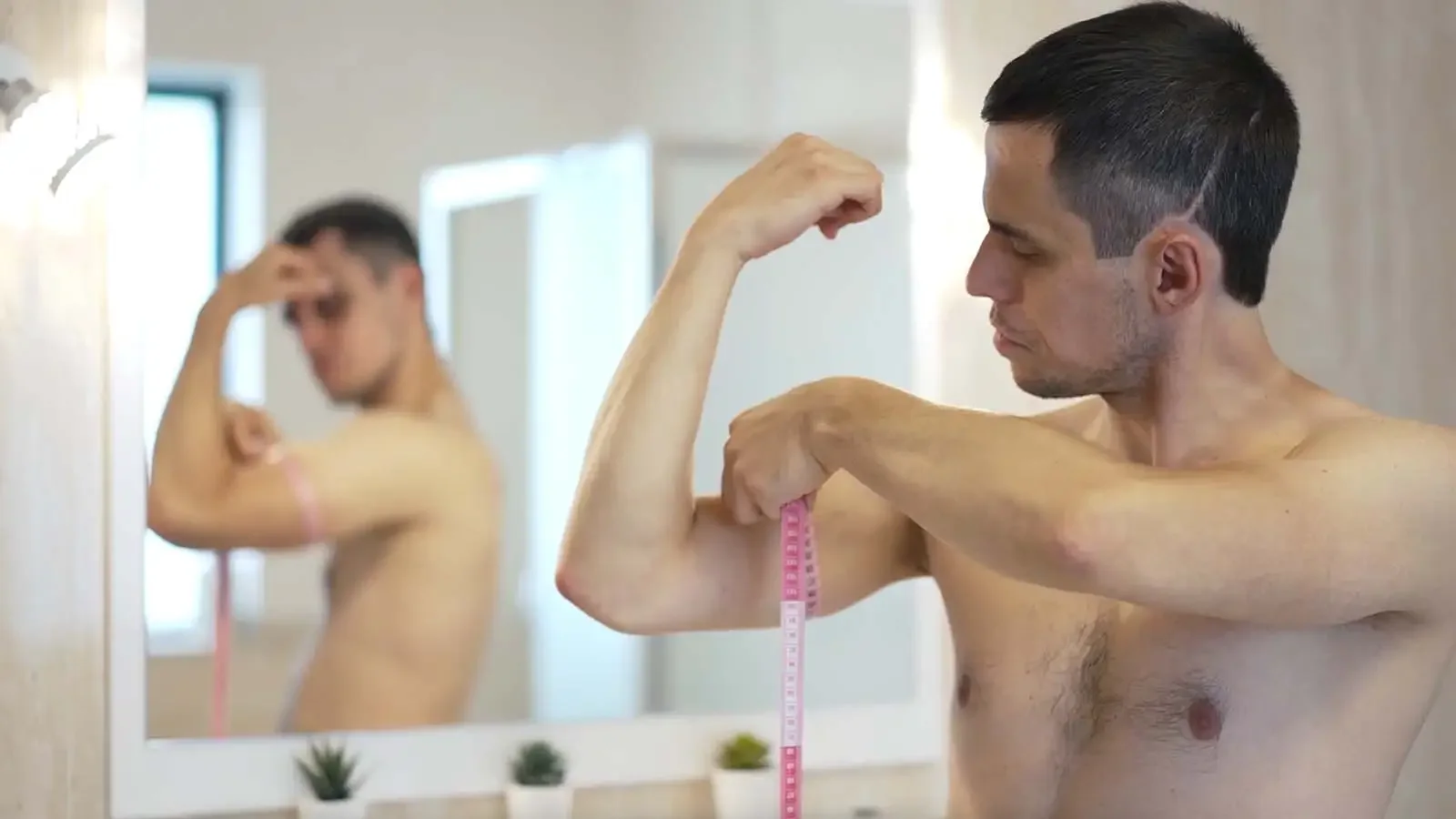 A shirtless man measuring his bicep with a pink measuring tape in front of a mirror, with his reflection also visible.