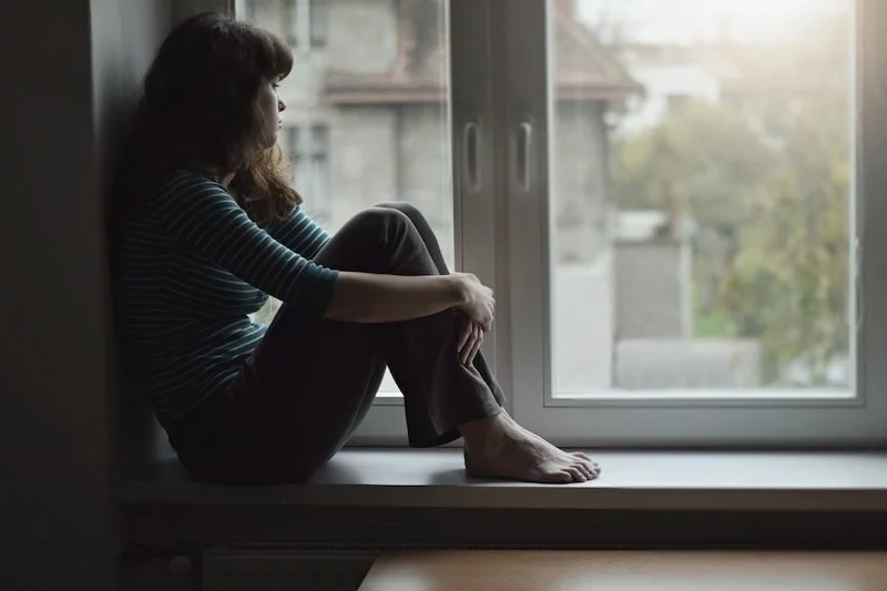 A woman sitting on a windowsill, looking out at a cloudy outdoor scene with trees and buildings, with a contemplative or sad expression.
