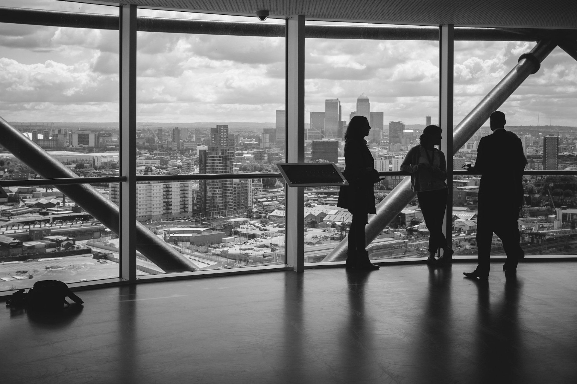 Silhouettes of three people standing inside a modern building with large windows, overlooking a city skyline with high-rise buildings and a partly cloudy sky.