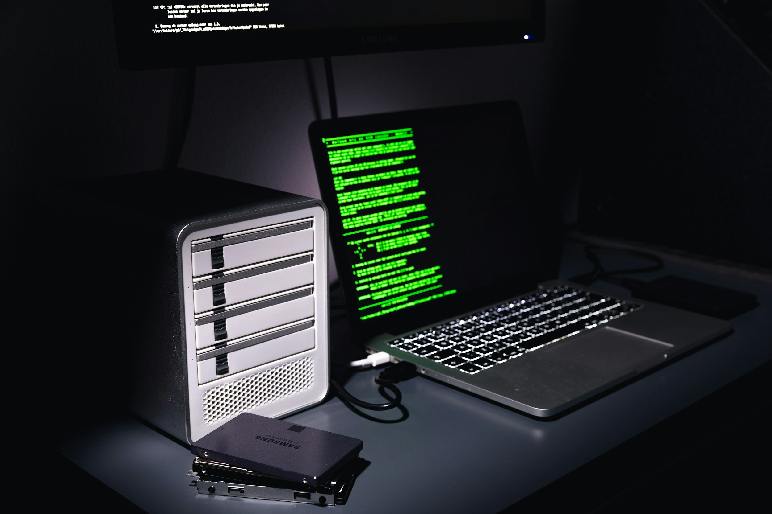 Dark workspace with a computer monitor displaying code, a laptop with code on its screen, a computer tower, and two Samsung solid-state drives arranged on a desk.