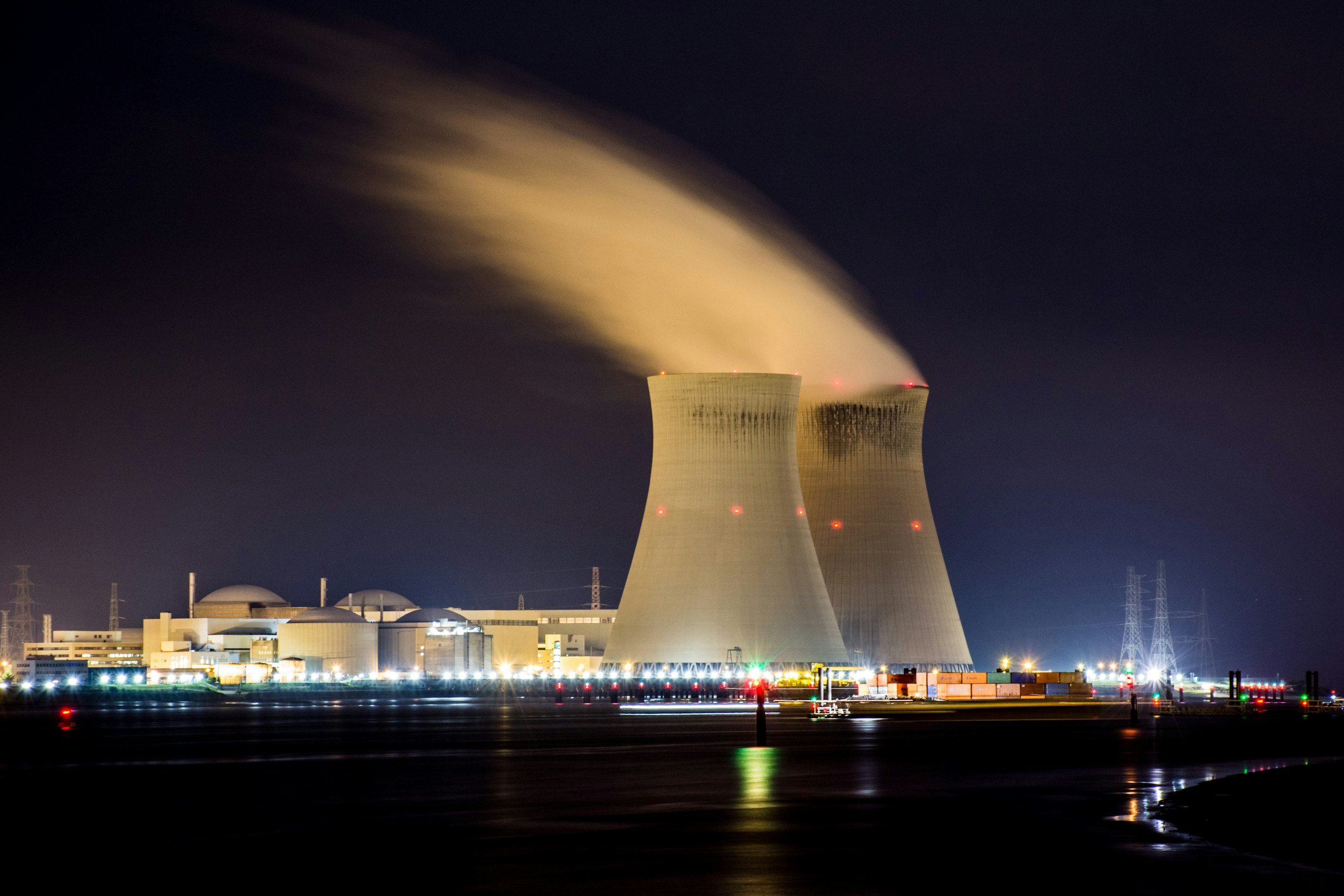 Nighttime scene of a nuclear power plant with cooling towers emitting steam, illuminated by exterior lighting, and shipping containers visible in the foreground.