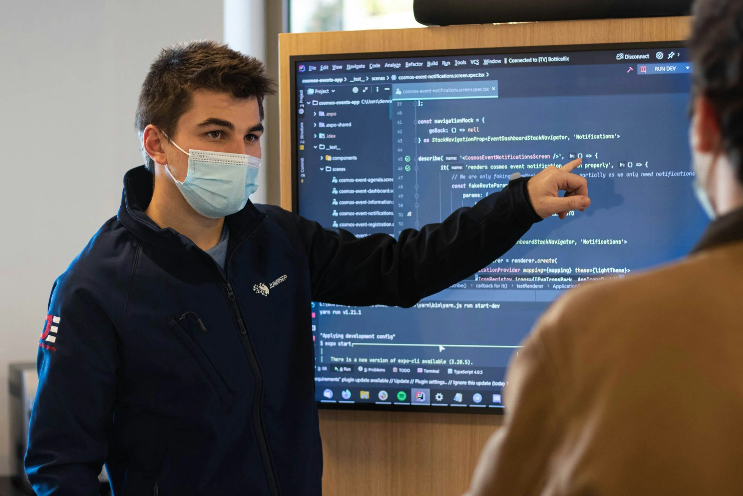Young man wearing a face mask and a dark jacket explains coding on a large monitor to another person, with a window in the background.