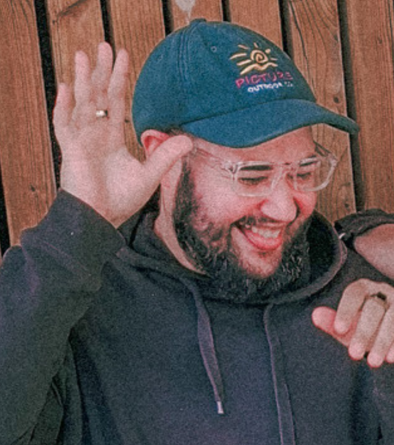 Un homme souriant, portant une casquette bleue avec un logo coloré, des lunettes transparentes, et une veste noire, gesticulant avec la main, devant une clôture en bois.