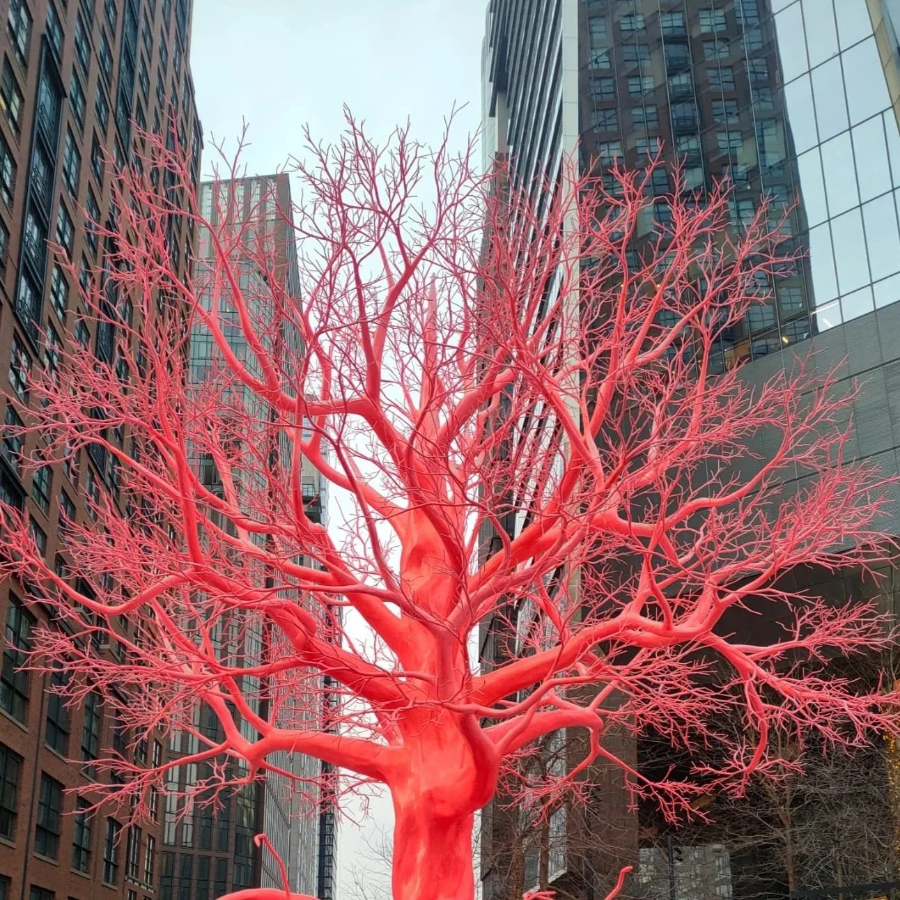 Arbre rouge artistique en plein air dans une zone urbaine, entouré de bâtiments modernes en verre et en brique.