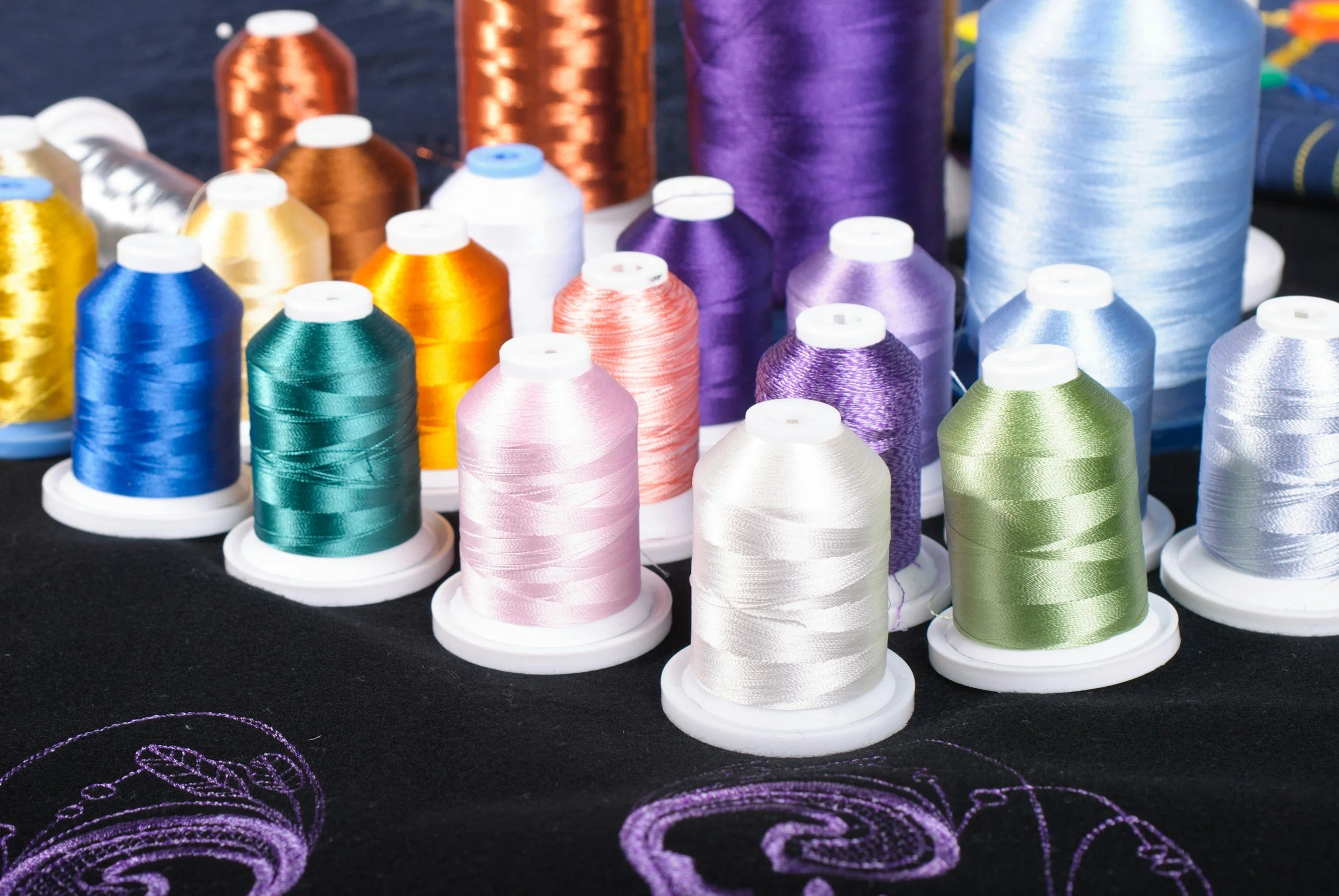Various spools of colorful embroidery thread including metallics in colors like purple, blue, green, pink, orange, gold, silver, and copper, placed on a black surface with purple embroidered thread patterns visible in the foreground.