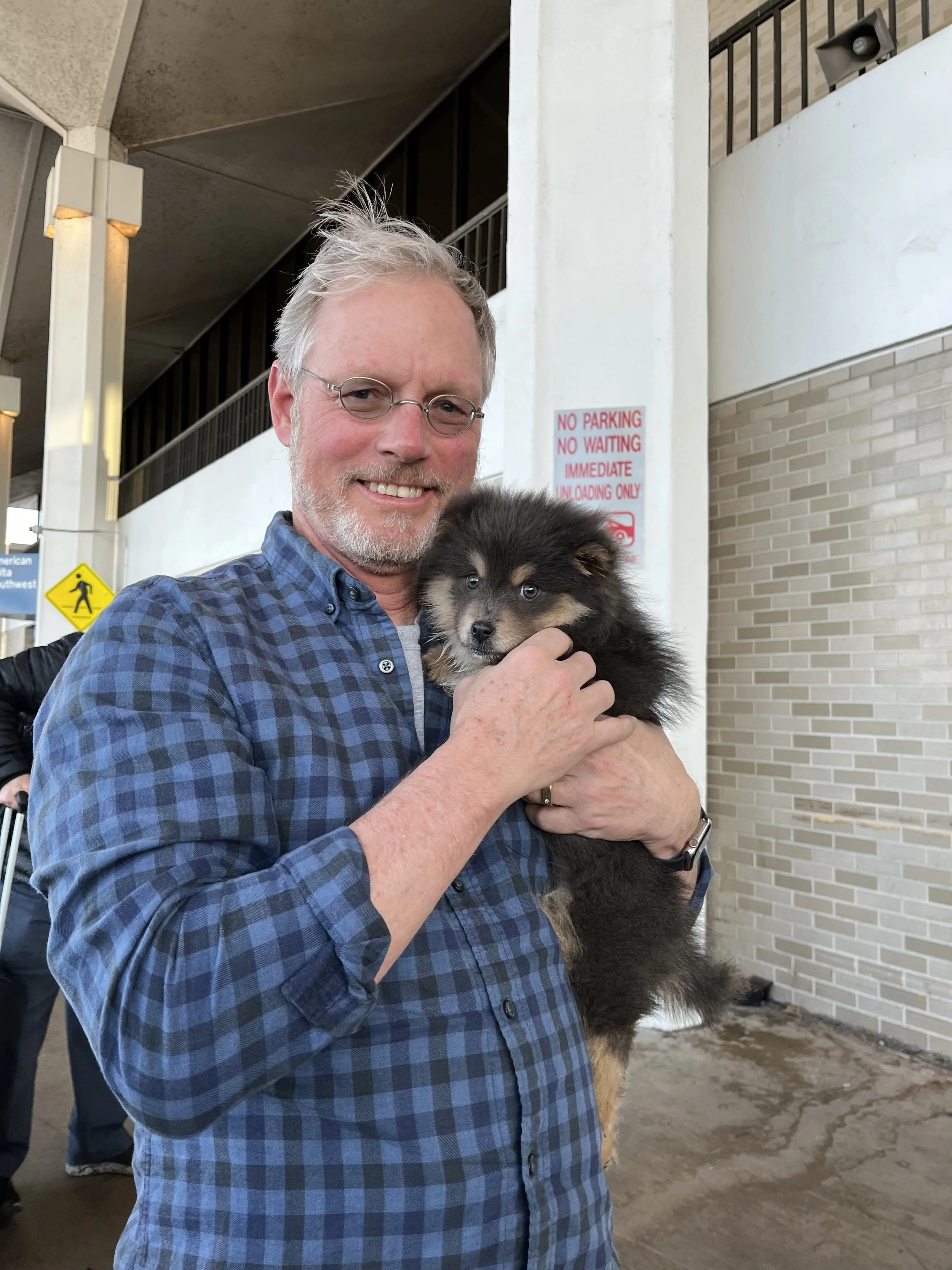 A man with glasses and a blue plaid shirt smiling while holding a black and tan puppy outdoors near a building. There are signs and a staircase in the background.