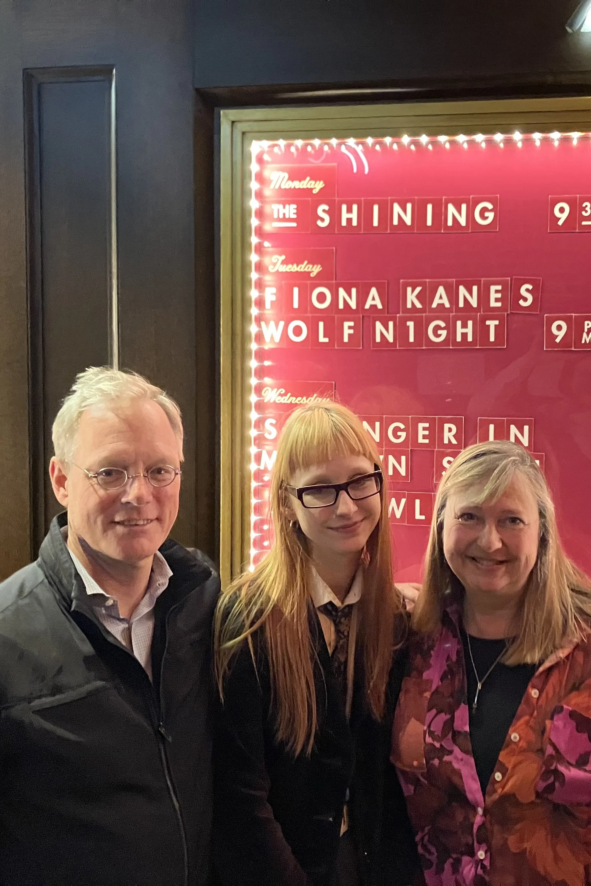 Three people standing close together in front of a red poster board with white text, in a dimly lit room. The poster lists days of the week and event titles, including 'The Shining' on Monday, 'Fiona Kanes' and 'Wolf Night' on Tuesday.