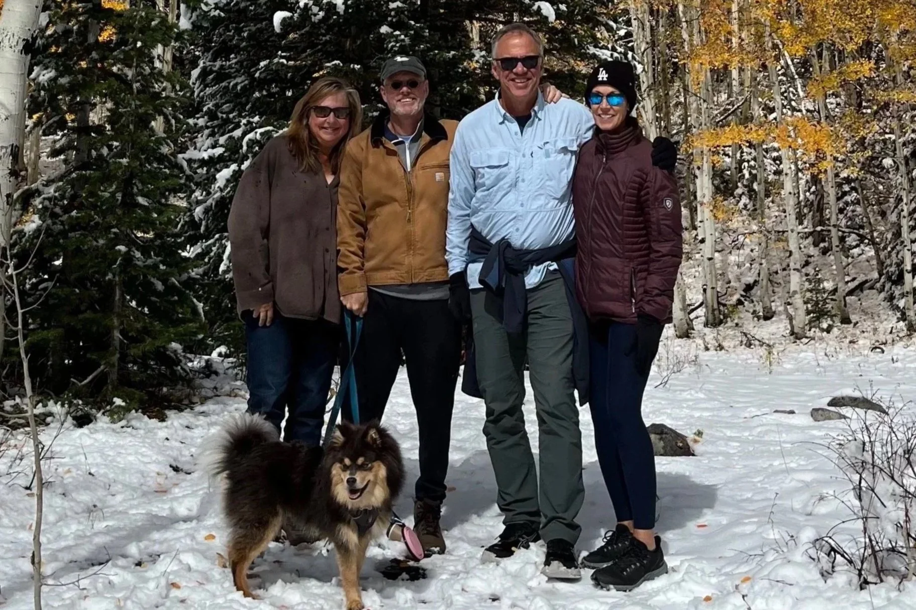 Four people standing together in a snowy forest, with a dog at their feet, smiling and wearing winter clothing.