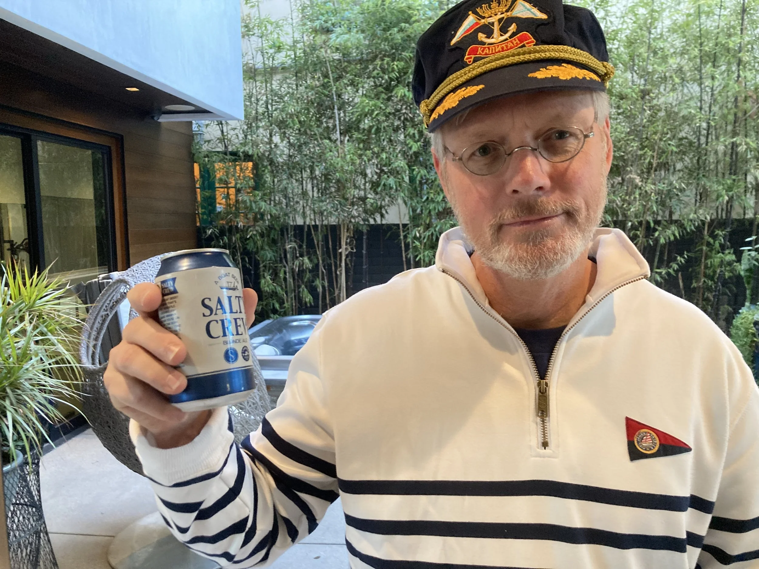 A man wearing glasses and a captain's hat with an anchor emblem, holding a salt cream container, sitting outdoors with greenery and a modern house in the background.