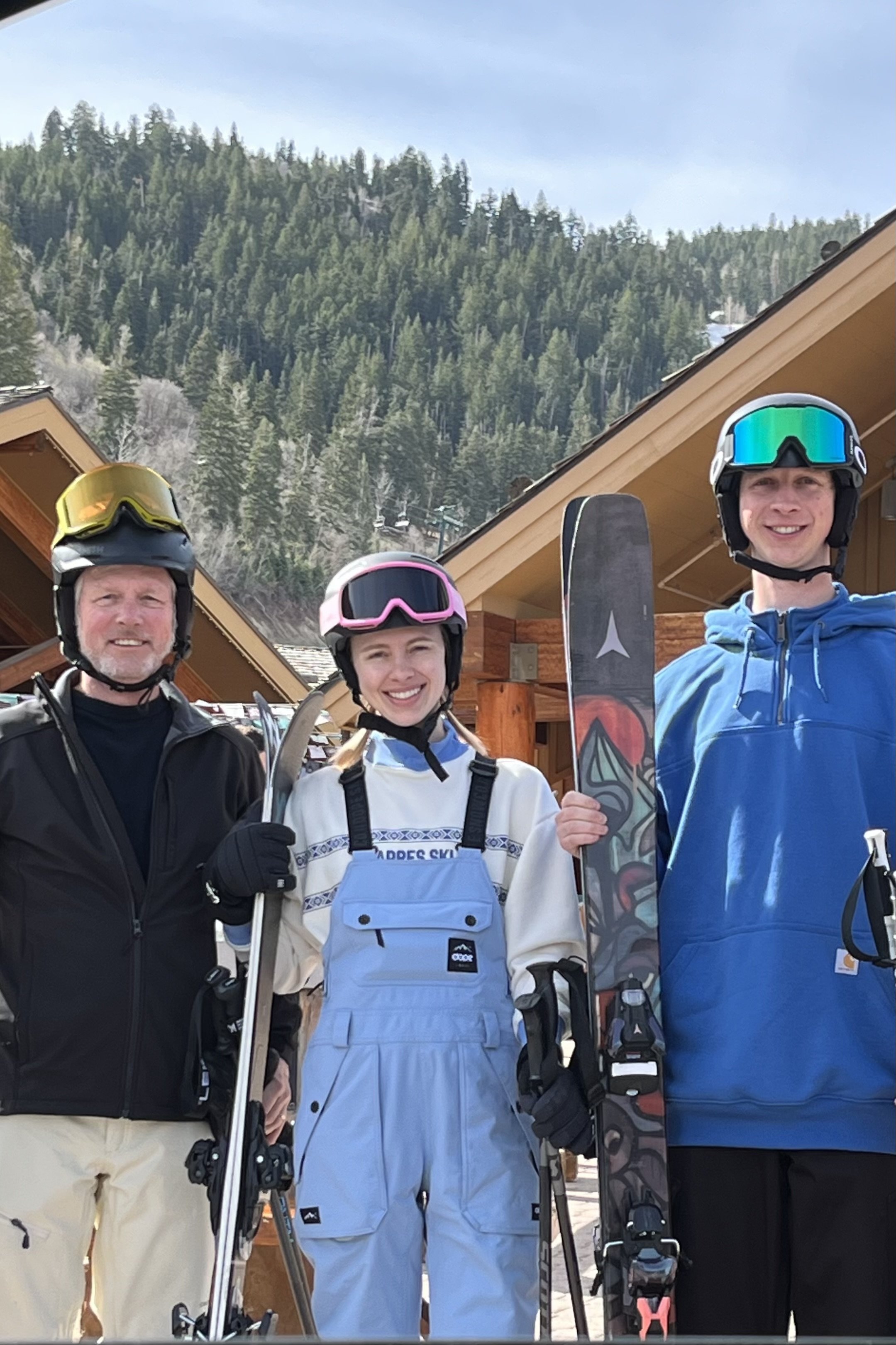 Three people dressed in ski gear standing outdoors in front of wooden buildings with a mountainous, forested landscape in the background. They are smiling and holding skis and poles.