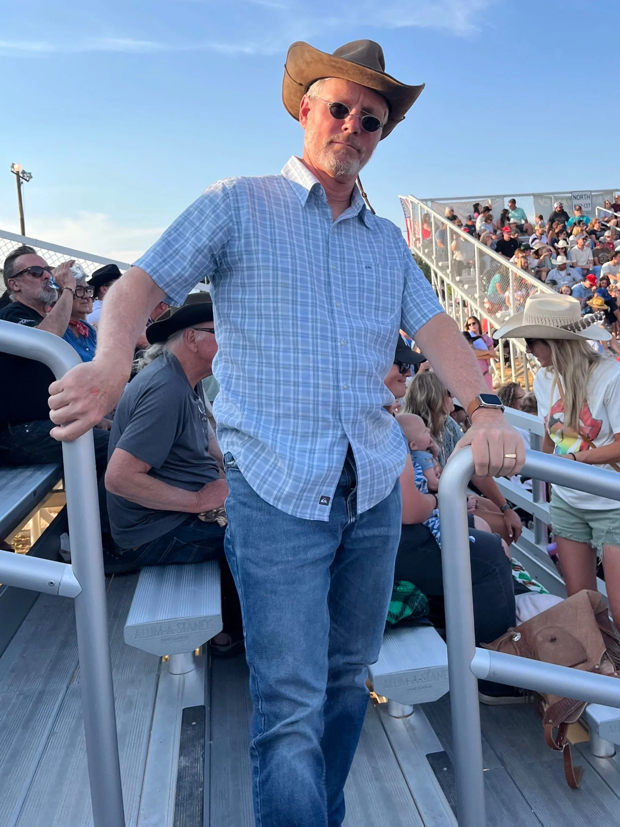 A middle-aged man wearing sunglasses, a cowboy hat, a short-sleeved plaid shirt, and jeans, standing on bleachers at a crowd-filled outdoor event, with other spectators seated behind him.