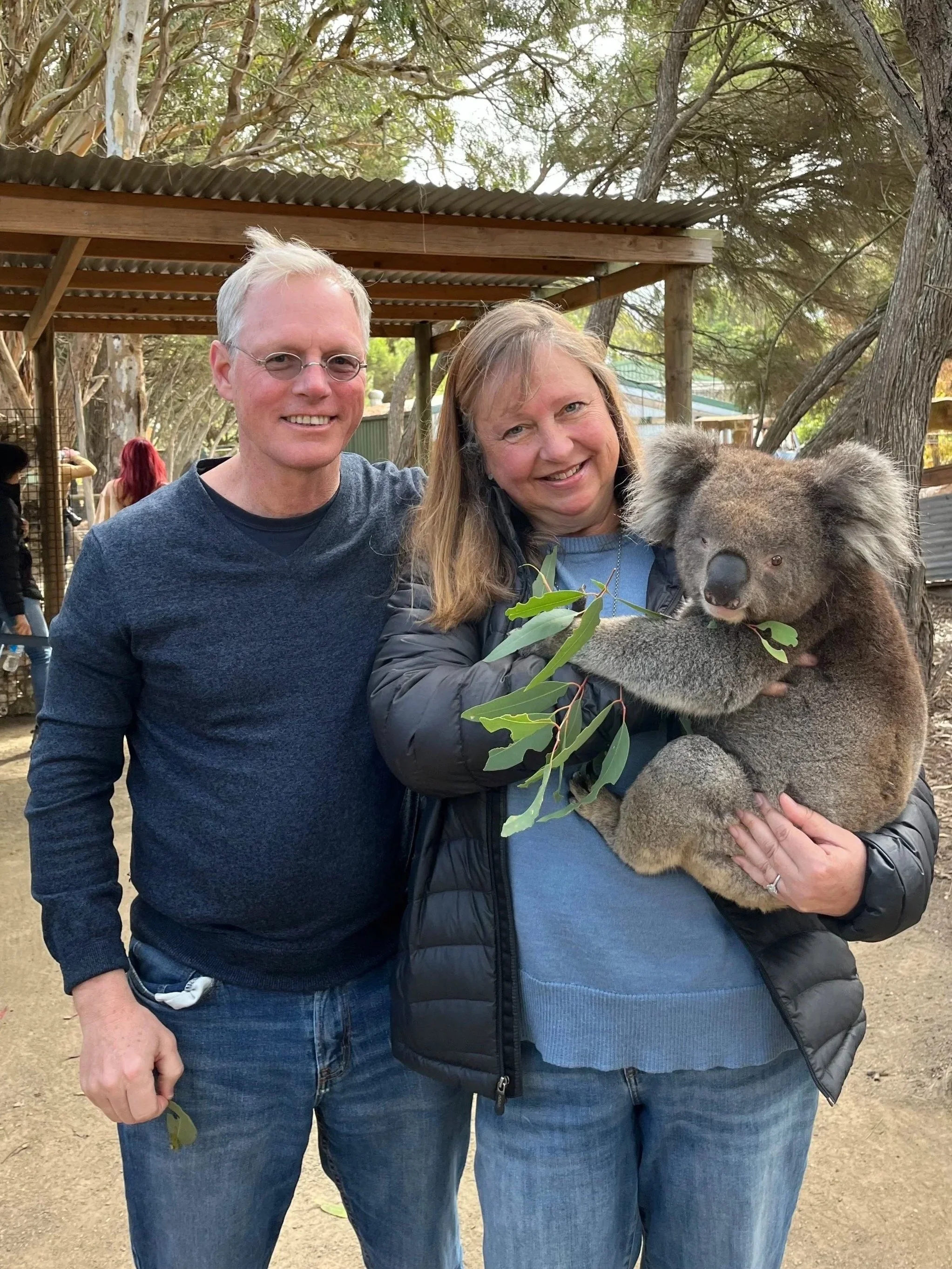 Two adults, a man and a woman, standing together outdoors at a zoo or wildlife sanctuary, holding a koala bear. The man has short gray hair, glasses, and is wearing a dark blue sweater. The woman has long light brown hair, and is wearing a dark jacket and blue shirt. The koala is gray with large ears, and is clutching some green leaves. Trees and a wooden shelter are visible in the background.