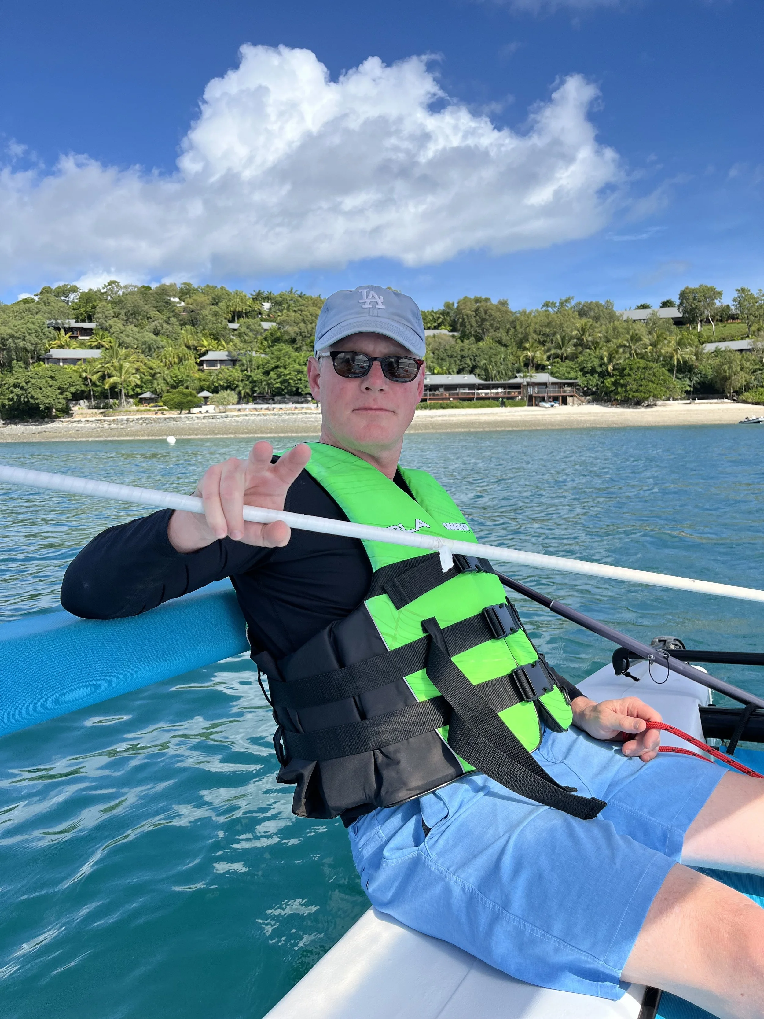 A man wearing a Los Angeles Dodgers cap, black sunglasses, a bright green life jacket, and blue shorts is sitting on a boat, holding a paddle, with a tropical shoreline of trees and buildings behind him. The sky is partly cloudy.