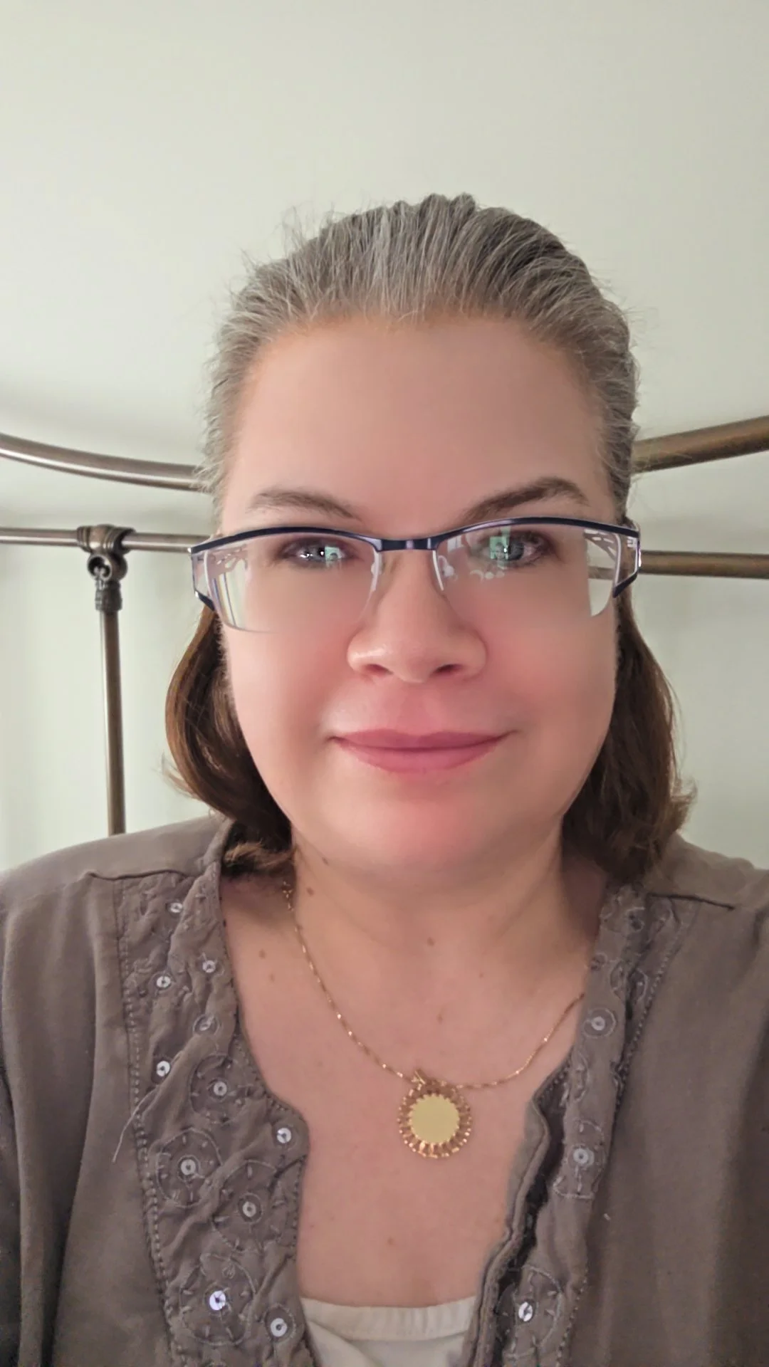 A woman with glasses, smiling, wearing a necklace, and a brown top with embroidery and sequins, in front of a metal bed frame.