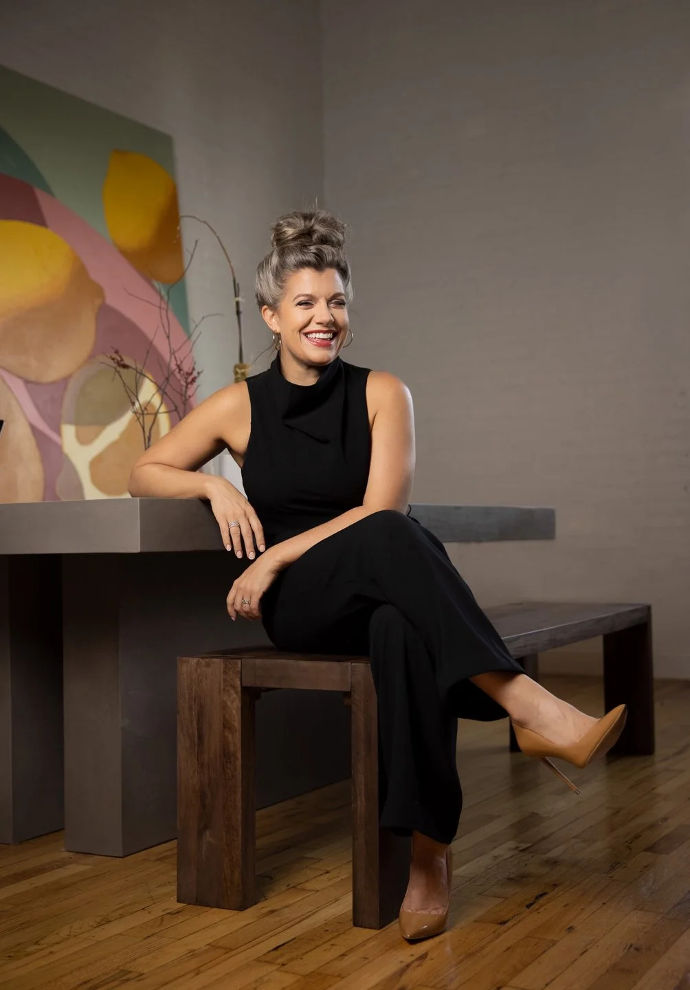 A smiling woman in a sleeveless black top and black pants sitting on a wooden bench, leaning on a wall with a colorful abstract painting, wearing nude high heels.