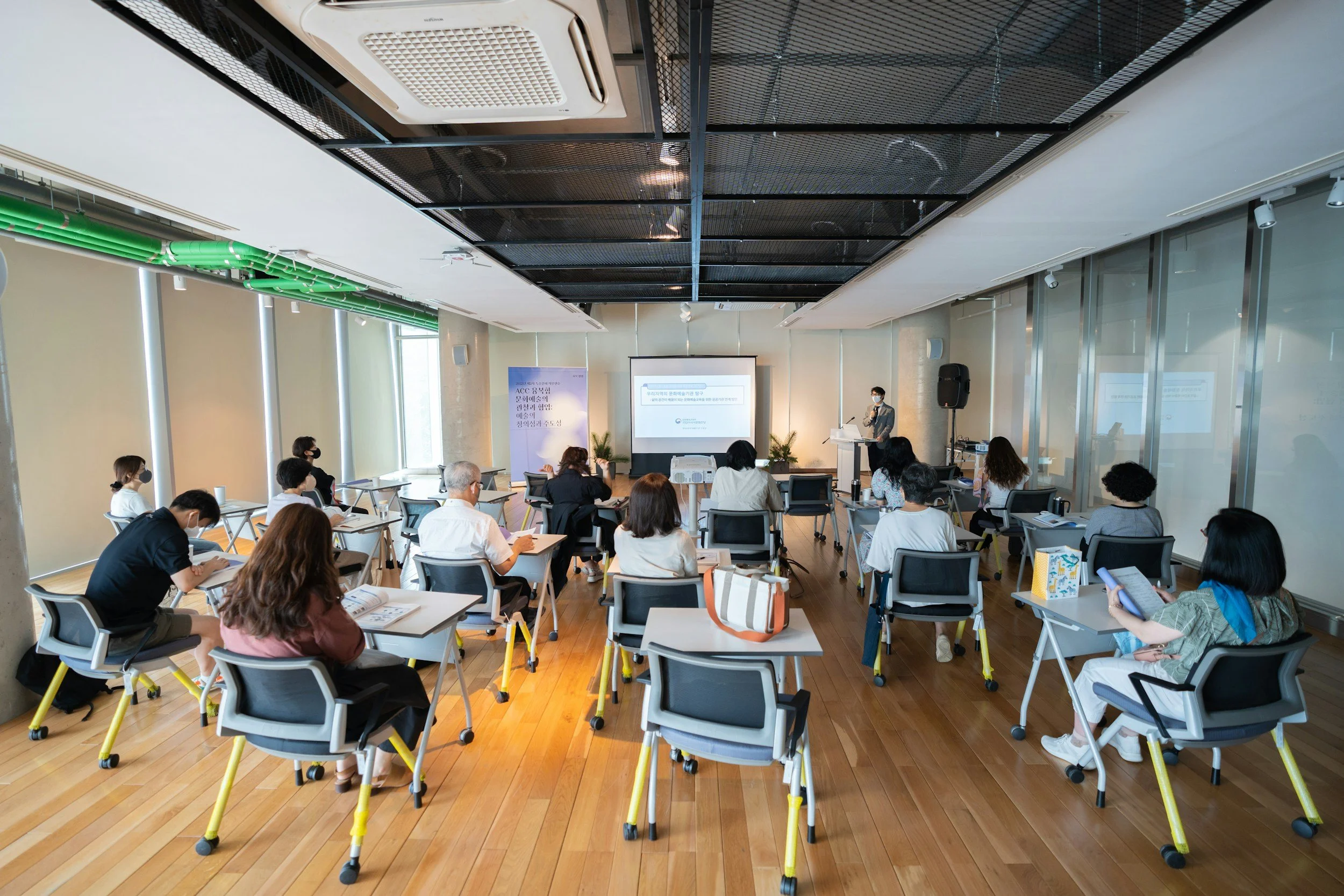A conference room with people attending a presentation. Participants are seated at tables, facing a speaker at a podium in front of a projection screen. The room has large windows with blinds, a wooden floor, and a black ceiling with exposed HVAC ducts and lighting.