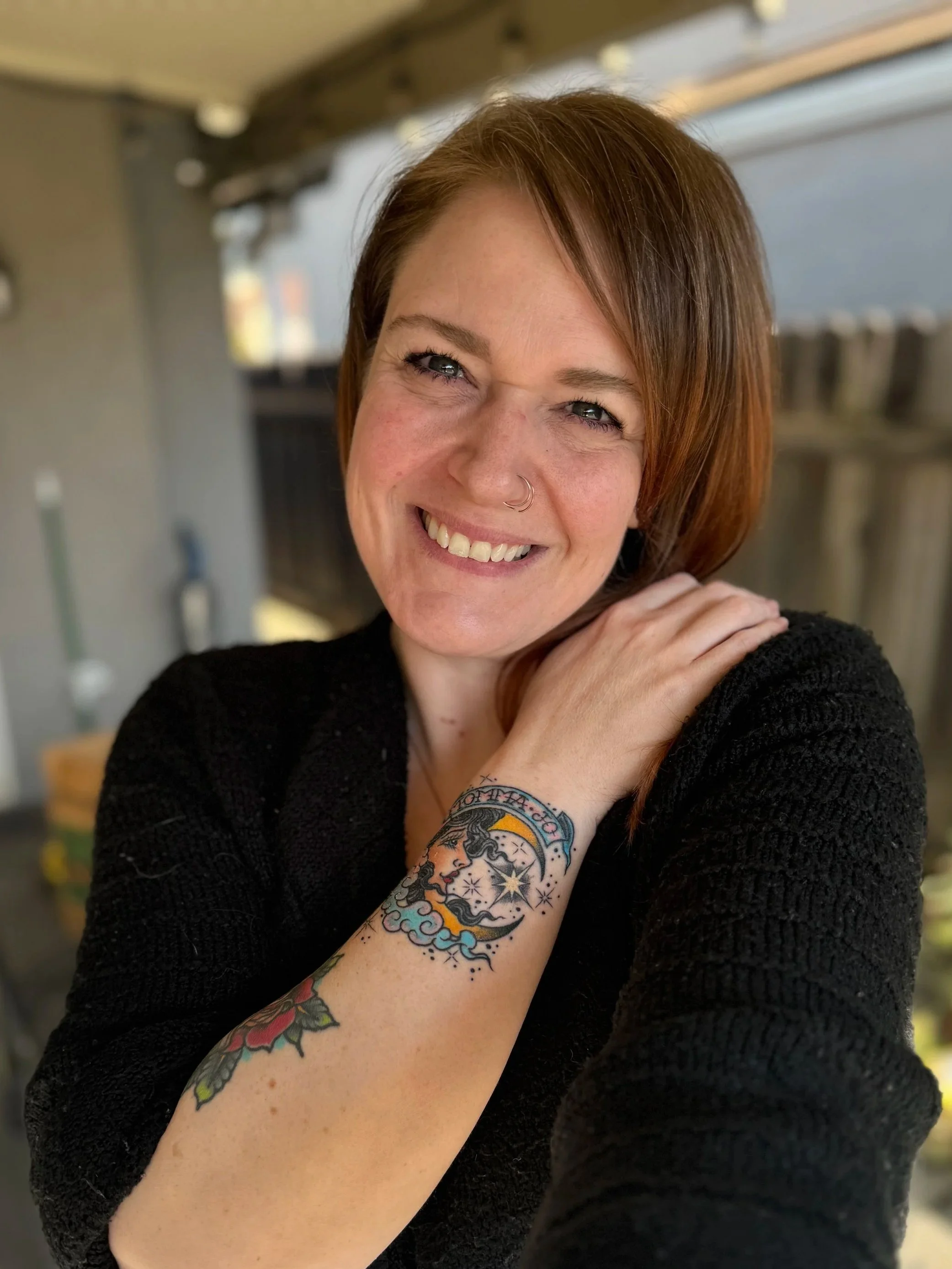 A woman with medium-length redish-brown hair smiling and showing her tattoos on her arm. Including a colorful American Traditional Style tattoo memorial saying Momma Jo using the trans flag colors. Wearing a black sweater outside.