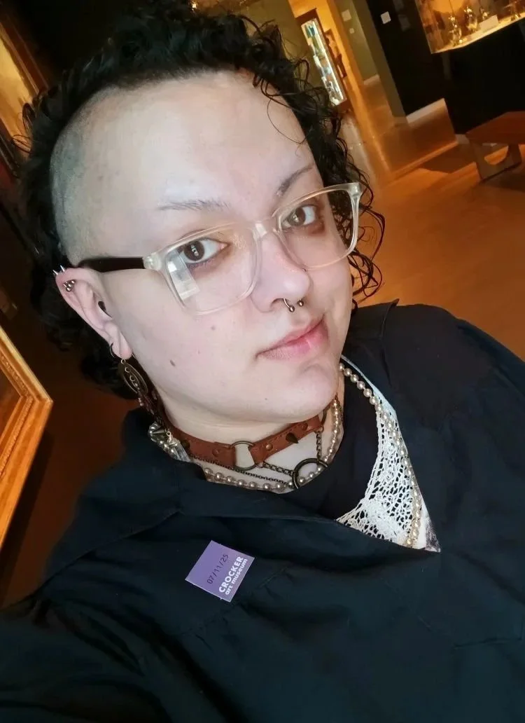 A person with short curly black hair, wearing clear glasses and multiple earrings, including a septum piercing, in a room with warm lighting and display cases of artifacts or collectibles in the background.