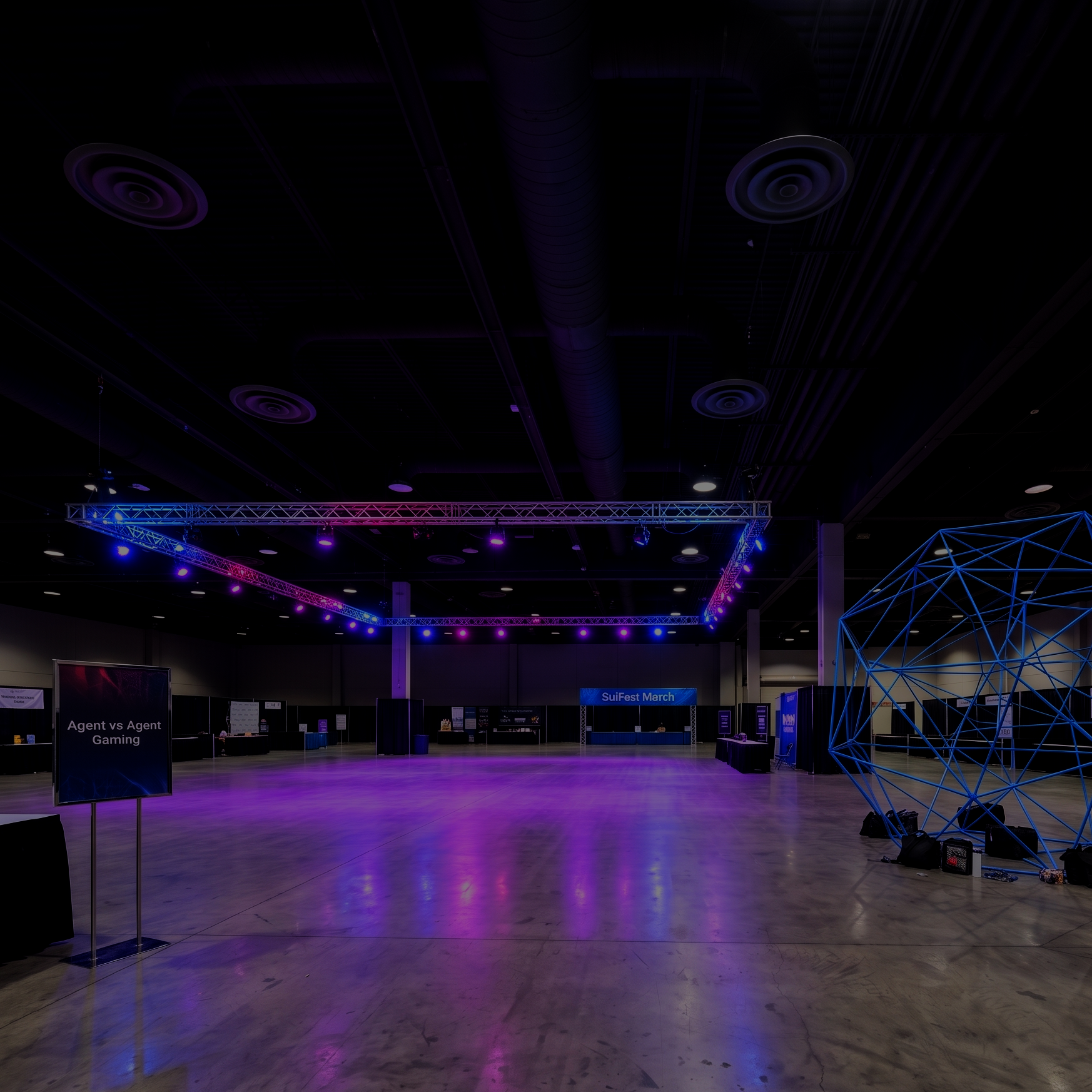 Wide view of an indoor event space with a wooden floor, illuminated by purple and pink stage lighting. There are display booths and signboards, including one that reads 'Agent vs Agent Gaming' and another that says 'SuiFest March.' On the right, there's a geometric wire sculpture illuminated with blue lights. The ceiling has dark panels with circular air vents and lighting fixtures.