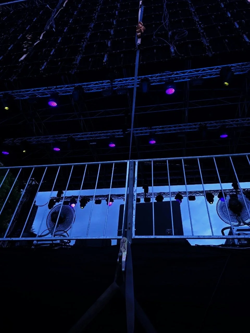 Stage with black metal framework, purple stage lights, and fans, viewed from below at dusk.