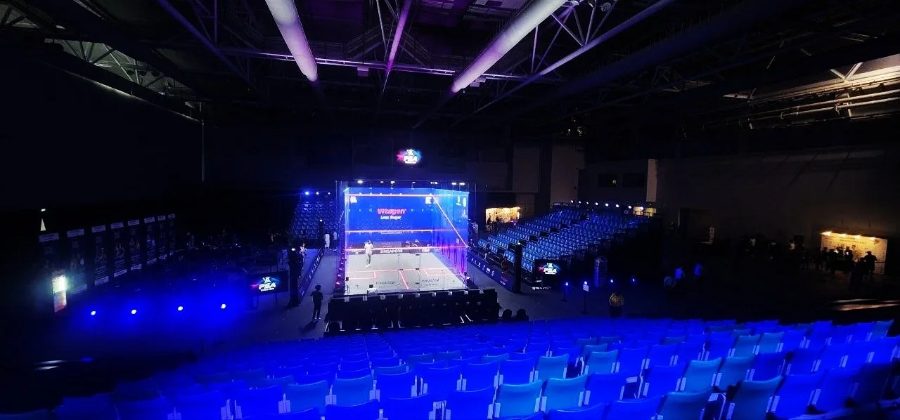 Empty indoor badminton arena with blue lighting, rows of seats, and a court in the center prepared for a match.