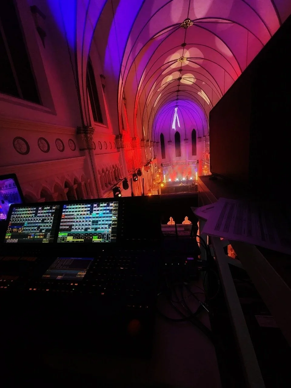 View of an indoor stage and event space with arched ceiling illuminated by colorful purple and red lights, seen from a sound and lighting control booth. The booth has digital equipment and papers.