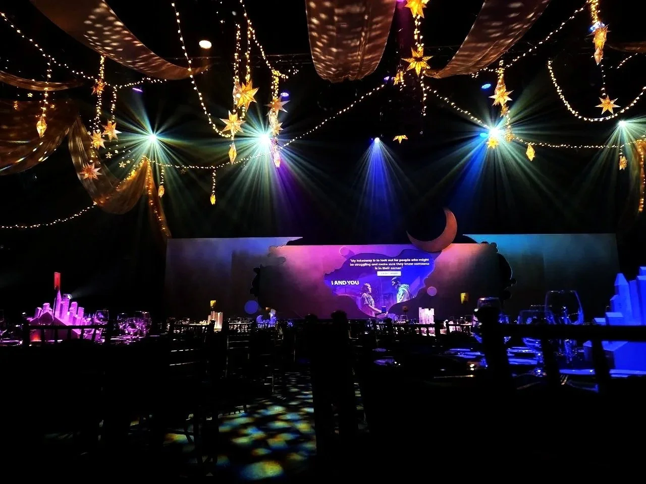 A dimly lit event space decorated with string lights, star-shaped lanterns, and hanging fabric, featuring a stage with vibrant colorful lighting, a crescent moon decoration, and a projection screen displaying text and images.