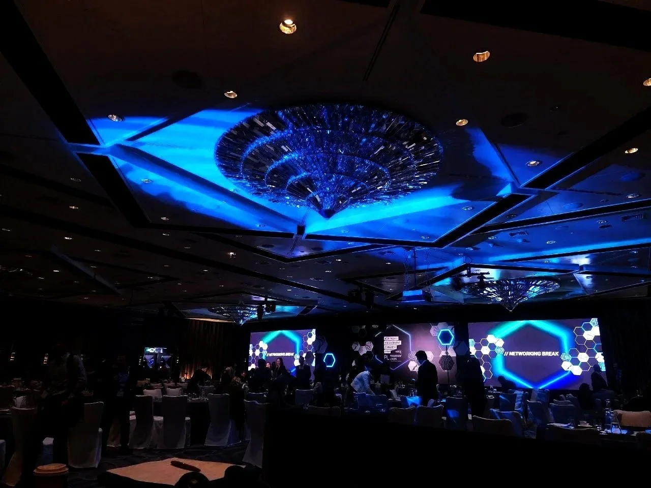 Dimly lit conference room with a large, illuminated blue chandelier on the ceiling, and multiple digital screens displaying a networking event.